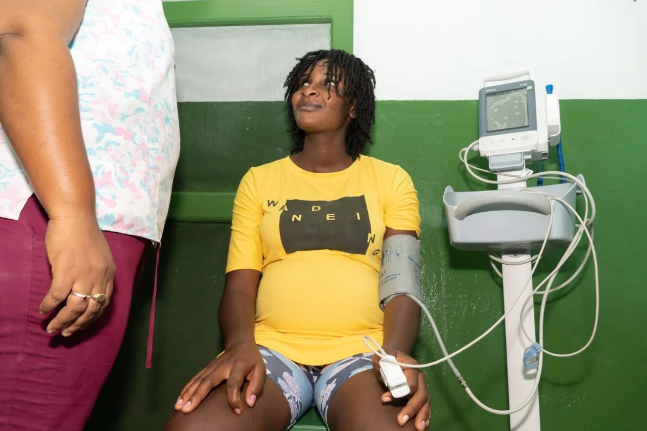 A pregnant woman sitting on a green bench in a medical room, with a blood pressure cuff on her arm and connected to a monitor, and a healthcare worker standing next to her.