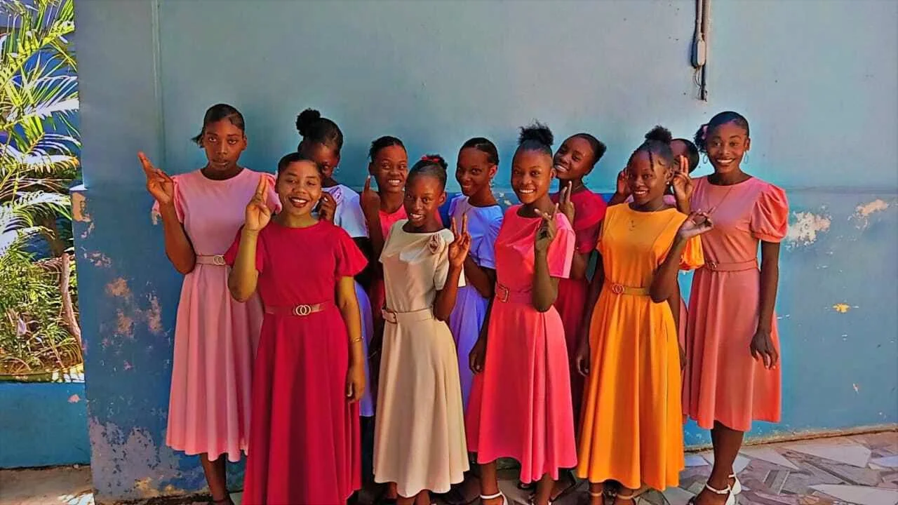 Group of ten young girls in colorful dresses standing in front of a blue wall, posing and smiling for the camera.