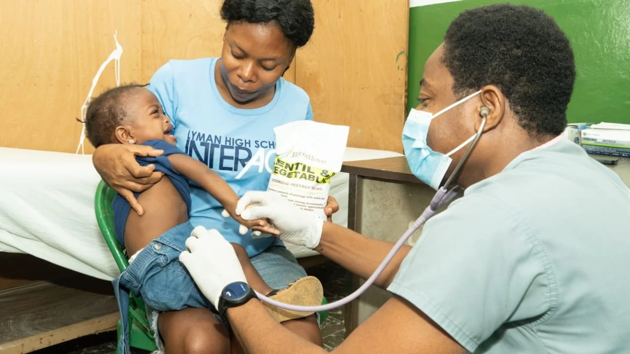 A healthcare professional wearing a mask and gloves examines a young boy who is sitting on a woman's lap in a medical clinic.
