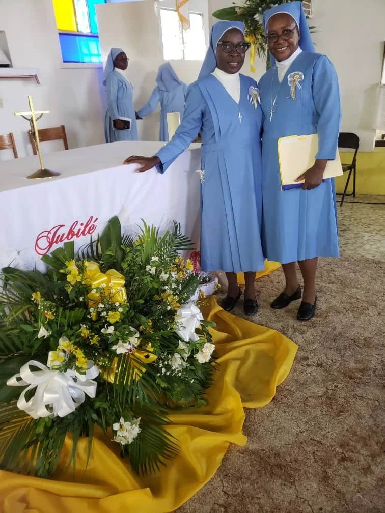 Two nuns in blue habits standing inside a church near a floral arrangement and an altar. One is holding a yellow folder, and there's a colorful stained glass window in the background.