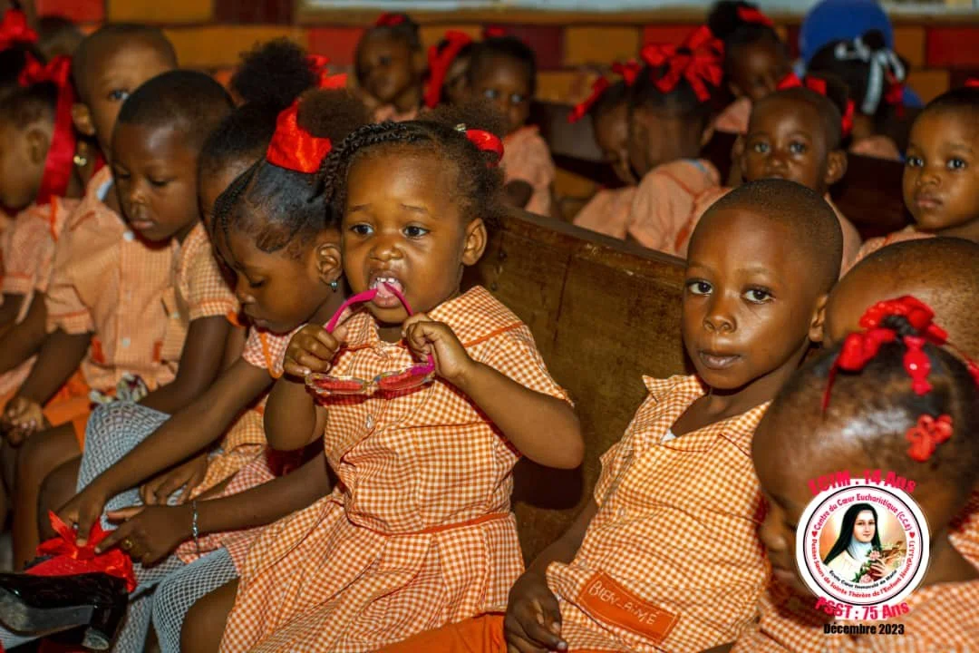 Group of young Haitian children sitting in a row on wooden benches, wearing orange checkered school uniforms, some with red bows in their hair, inside a classroom.