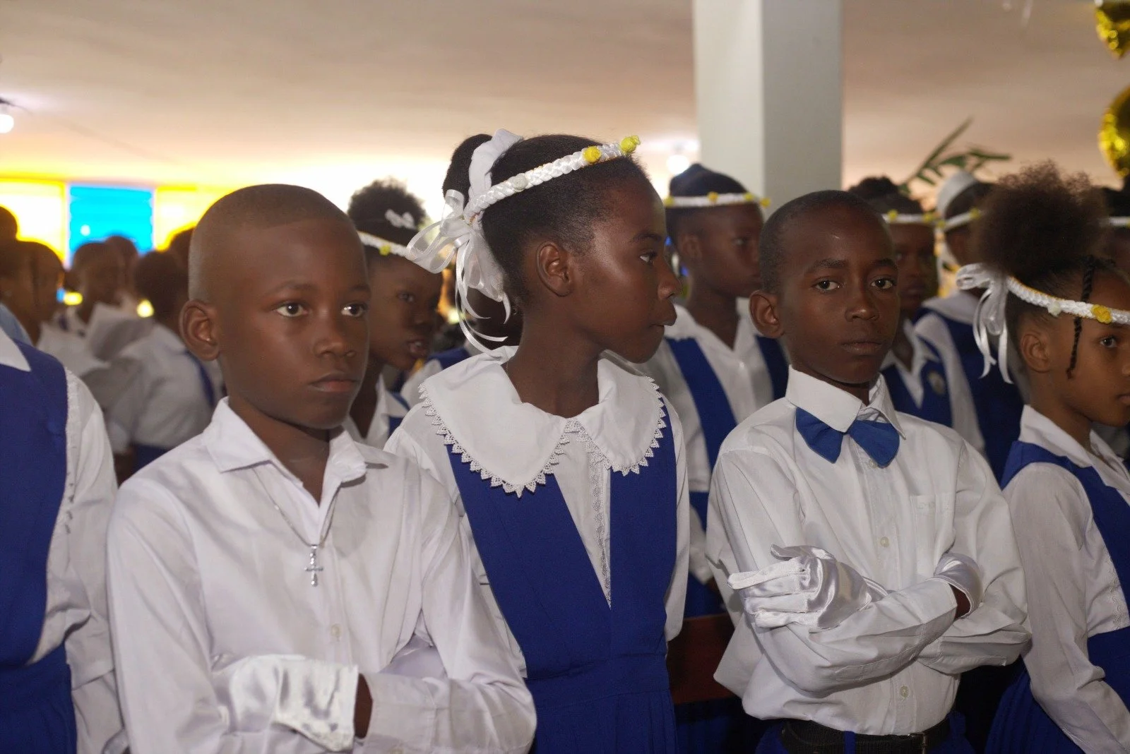 School children in white shirts and blue dresses or vests, many wearing gloves, attending a ceremony or event indoors.
