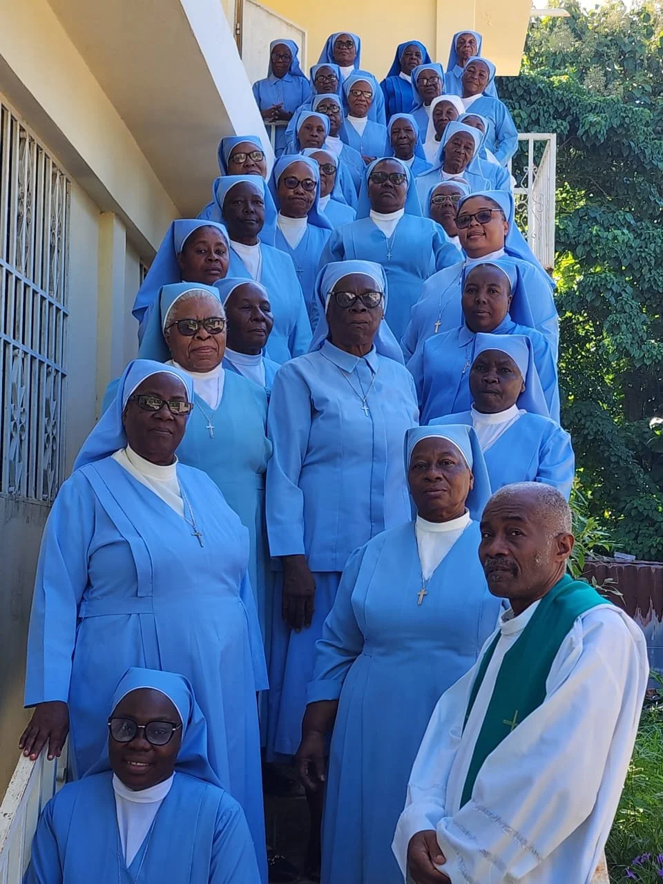 Group of nuns in light blue and white habit standing on stairs outside a building, with greenery in the background.