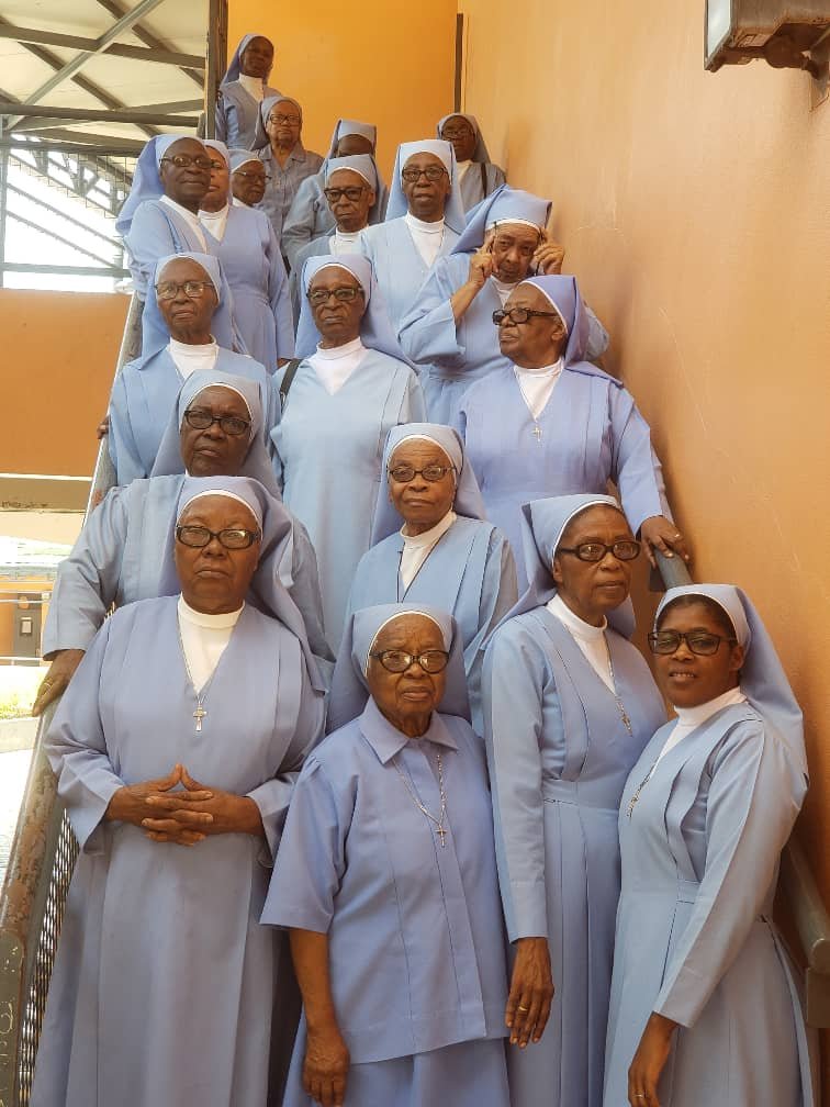 Group of Catholic nuns in light blue habits standing on a staircase, some wearing glasses, indoors with a yellow wall and a building outside visible.