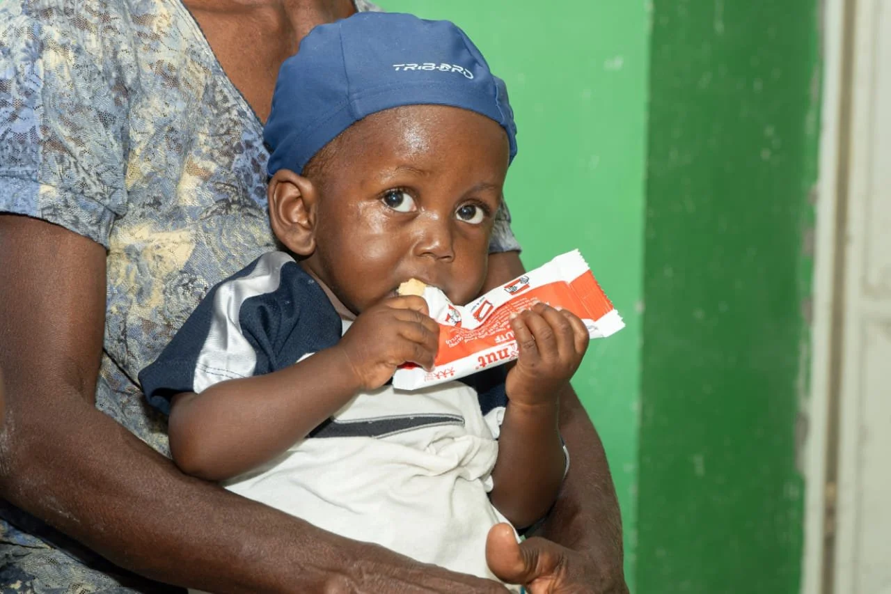 A young boy being held by an adult, wearing a blue cap, eating a snack from a red and white wrapper, with a green background.