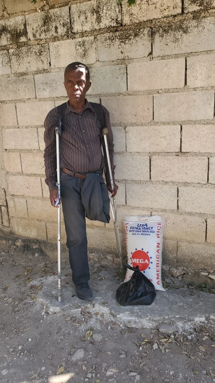 A man with crutches standing next to a bag of American Long Grain Rice against a concrete brick wall.