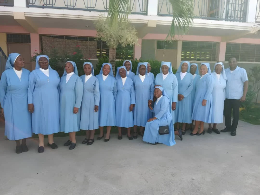 Group of women dressed in light blue religious habits, standing outdoors in front of a building, with one man in light blue shirt.