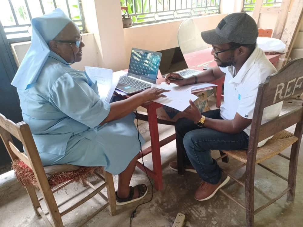 A nun in blue scrubs and a cap sitting at a table with a young man in a cap, glasses, and white shirt. They are discussing documents with laptops on the table, in a room with a window and natural light.