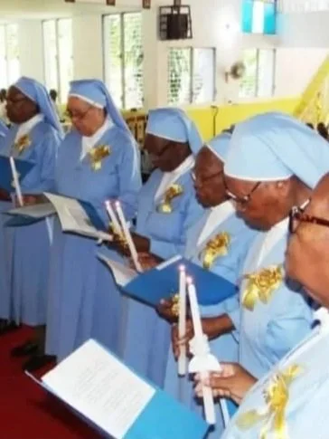 A group of women dressed in light blue religious robes and head coverings, holding candles and songbooks, in a church setting.