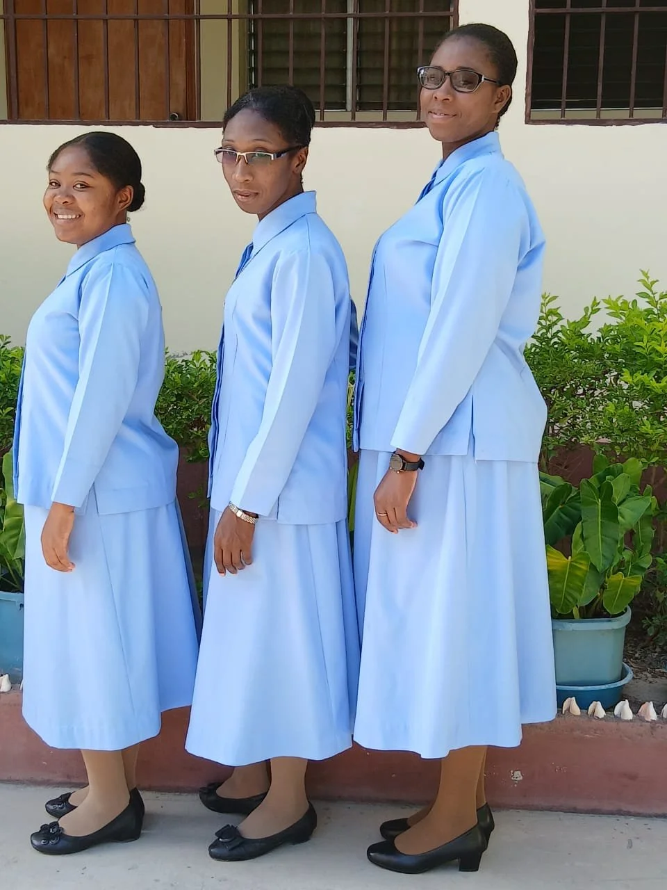 Three Haitian women in matching light blue uniforms with long skirts, black shoes, and glasses stand in a line outdoors, smiling for the photo.