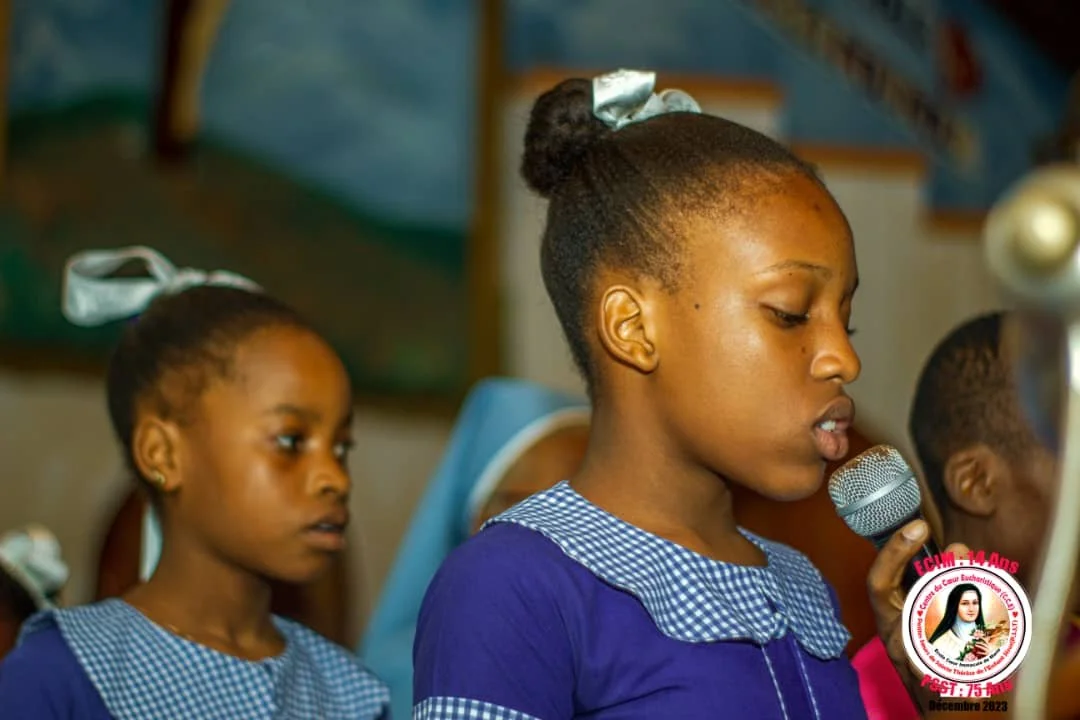 A young girl in a blue and white school uniform is speaking into a microphone, with another girl in similar uniform standing behind her, in a classroom setting.