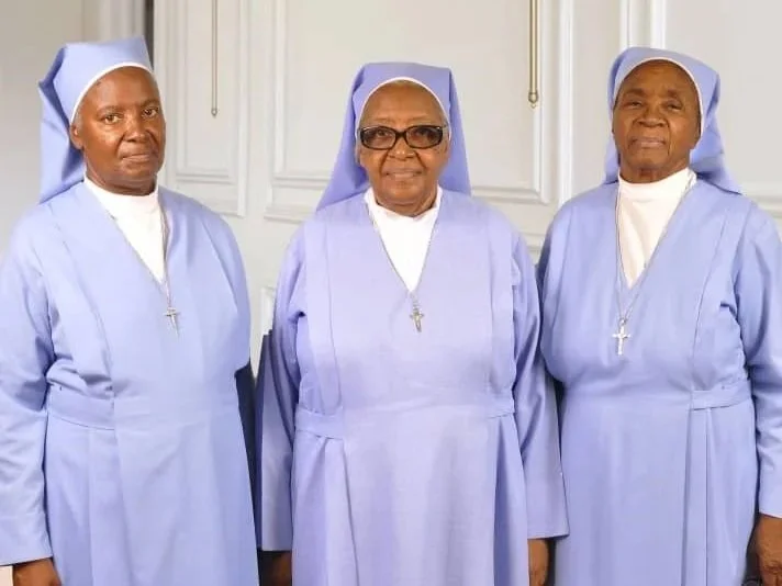 Three nuns standing together, dressed in light blue habits and wearing crosses around their necks, indoors with white walls.