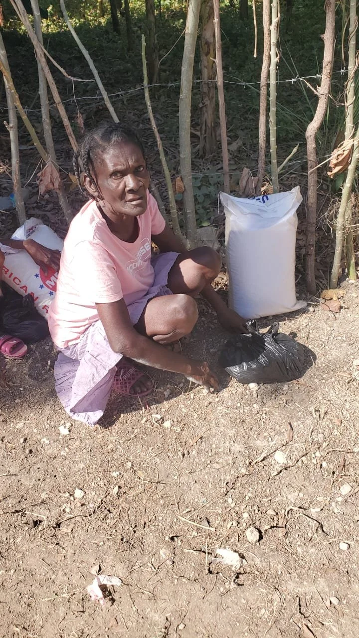 A woman receiving Non perishable dry food product from PSST Food Security Initiative, with large bags of supplies nearby.