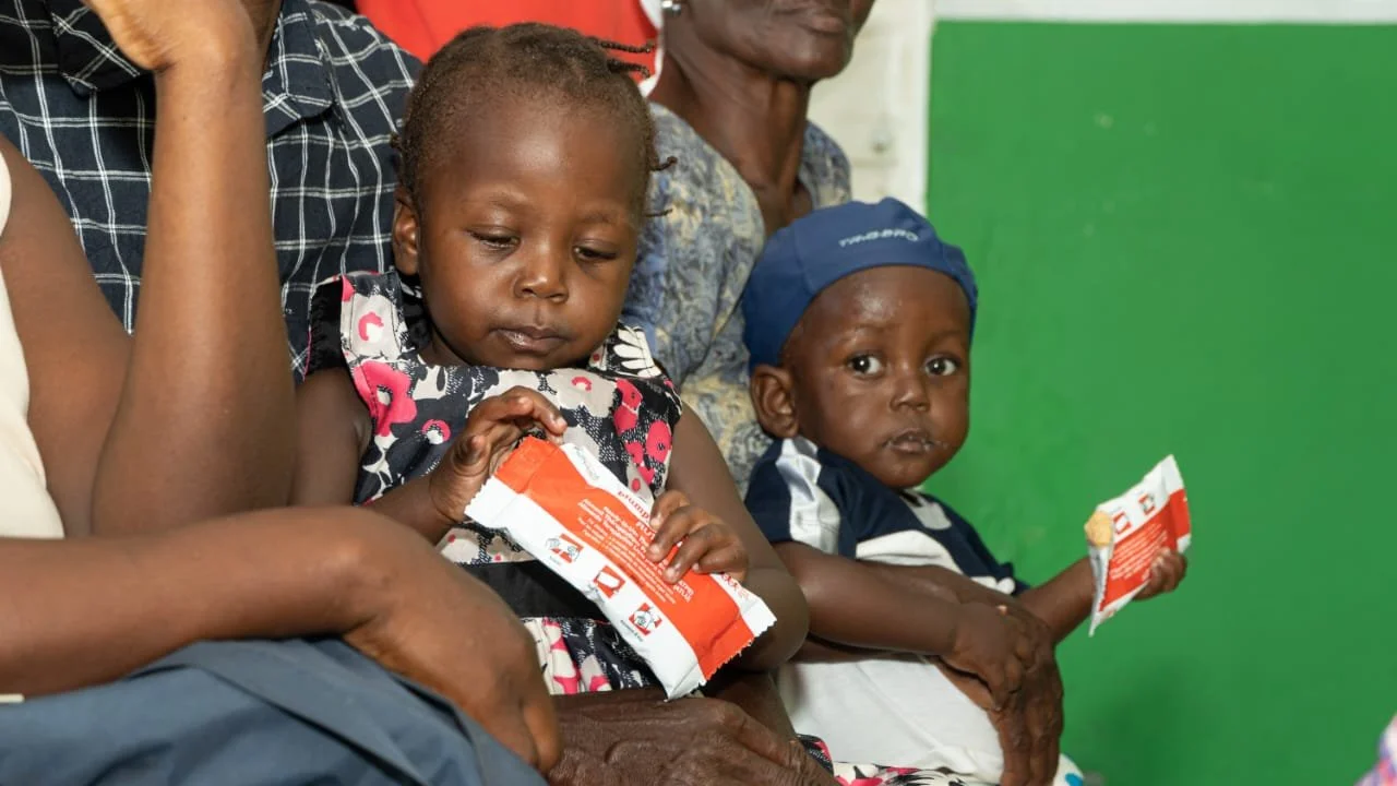 Two young children sitting on a woman's lap, holding snack packages, with an adult woman and a man in the background, in a room with green walls.