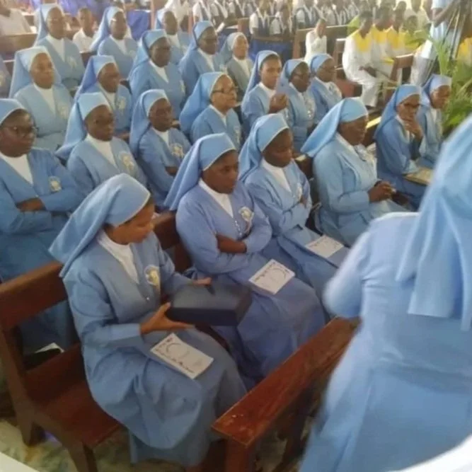 Group of nuns dressed in blue habits attending a religious ceremony in a church, with some holding books. The background shows additional people and religious decorations.