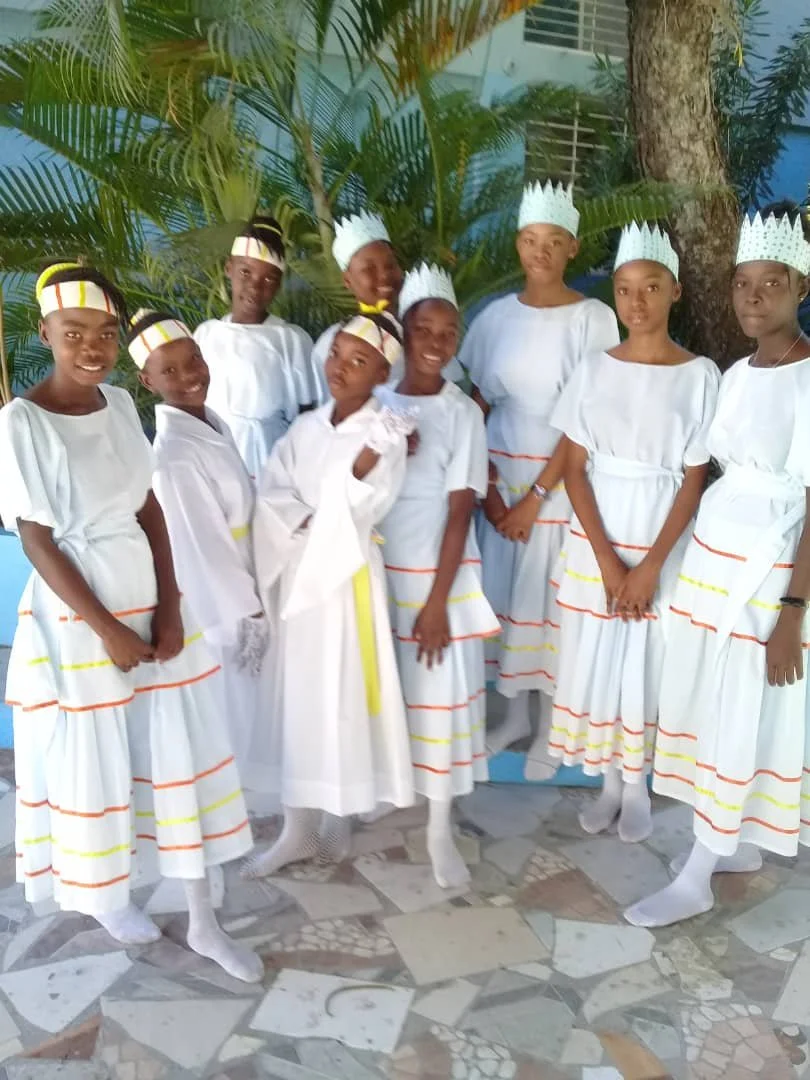 Group of young girls dressed in white costumes with colorful stripes, wearing paper crowns, standing outdoors in front of greenery.