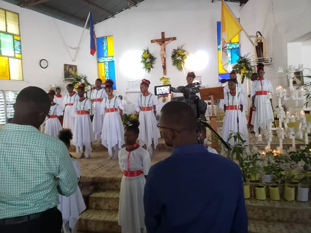 Children in white dresses with red belts standing in front of an altar with a crucifix, candles, and religious statues inside a PSST church.