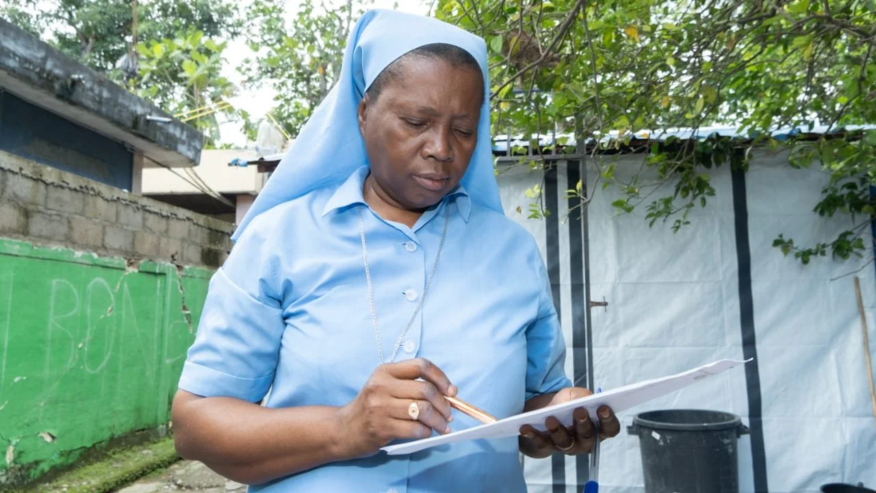 A woman dressed as a nun in a blue uniform with a blue head covering, holding and writing on a clipboard outdoors.