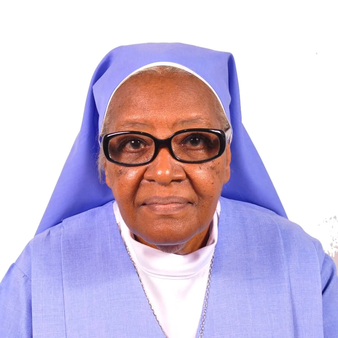 Close-up of an elderly woman nun wearing glasses, a blue habit, and a white head covering, sitting against a plain white background.