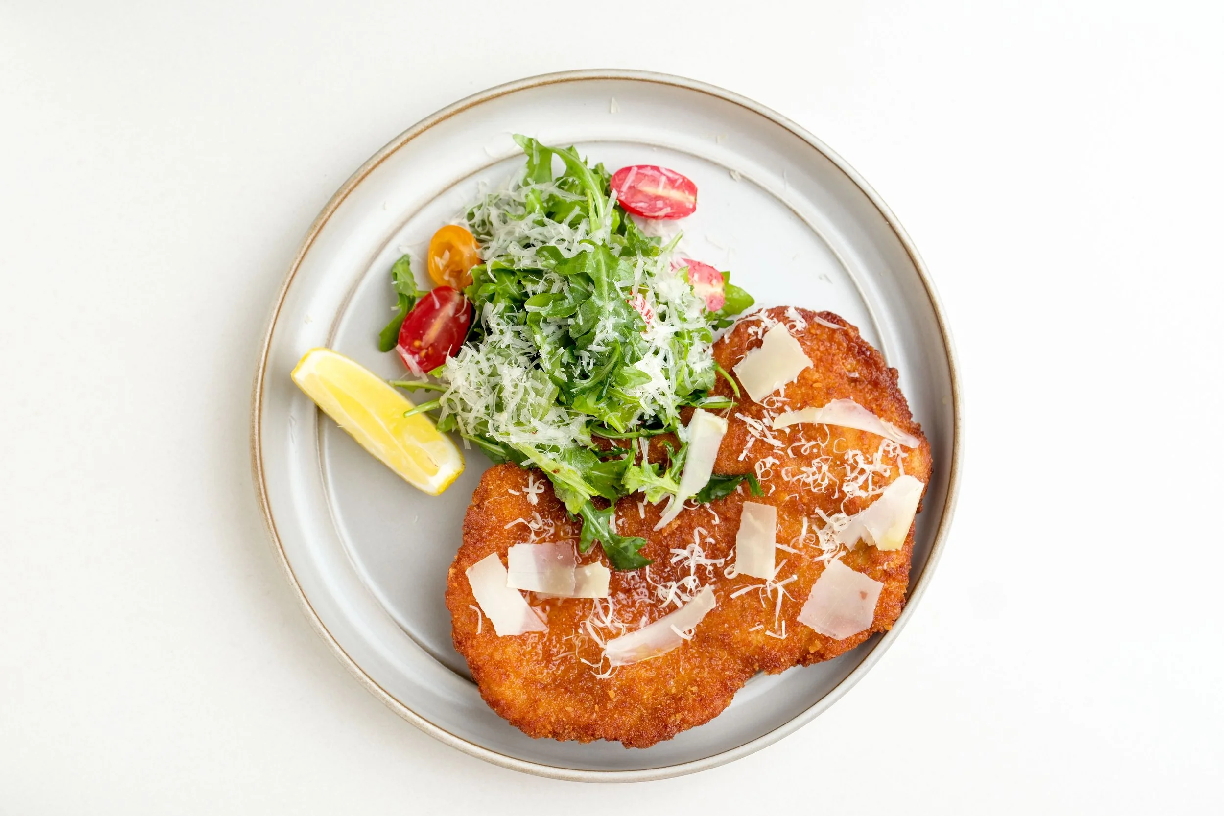 Fried breaded veal cutlet topped with shredded cheese, served with a fresh side salad including cherry tomatoes, arugula, and shredded cheese, with a lemon wedge on a white plate.