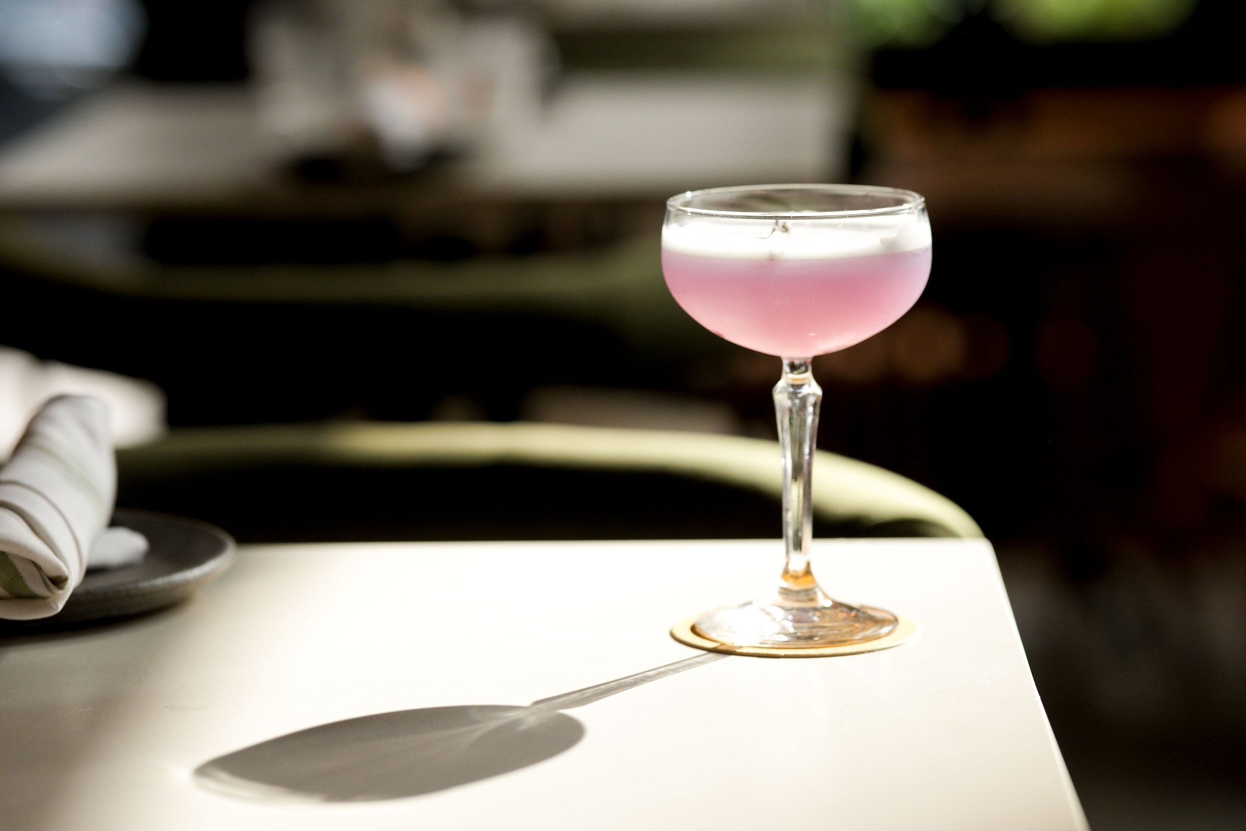 A pink cocktail in a coupe glass with a white foamy top, placed on a white surface, with a blurred background.