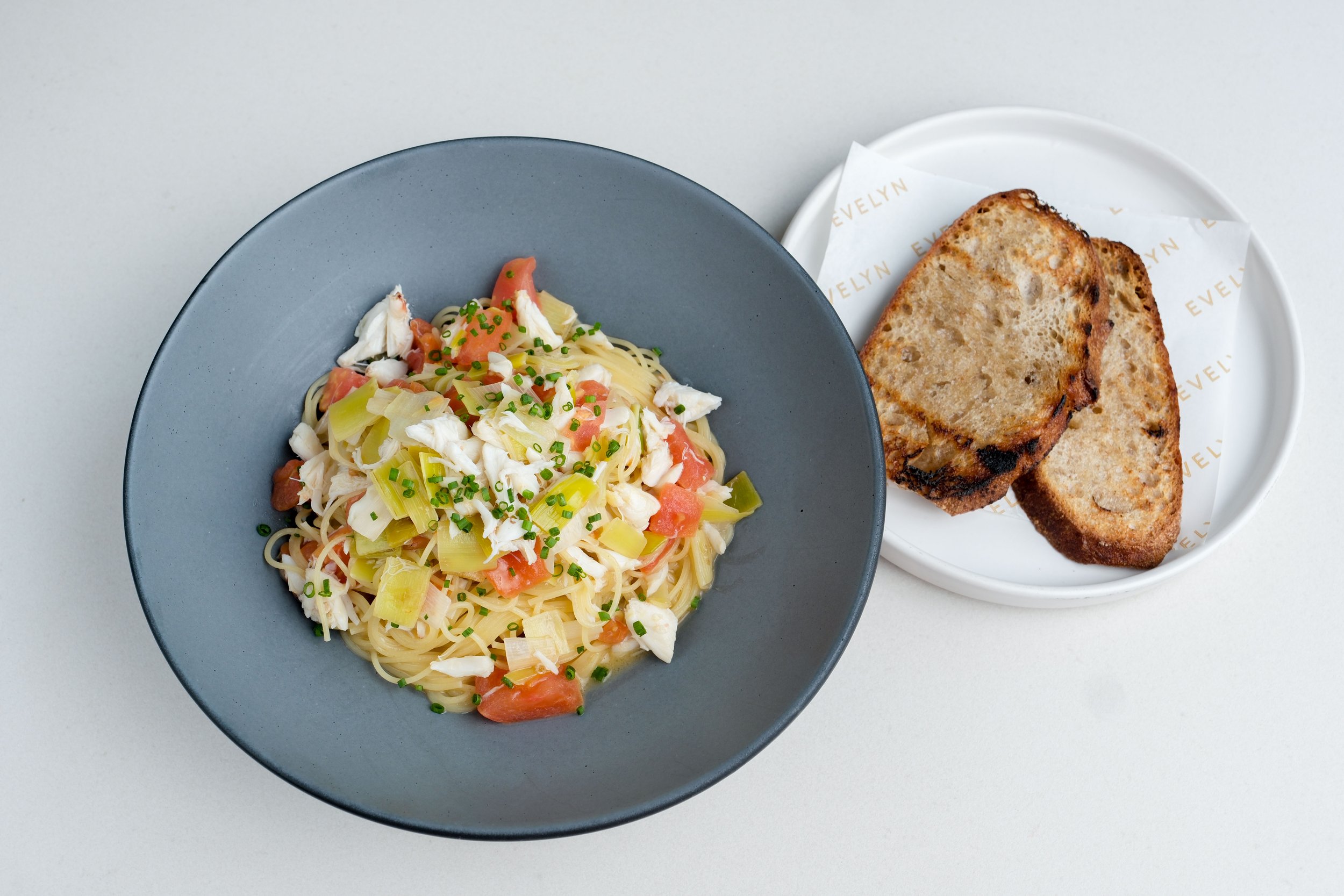 Bowl of spaghetti with chopped green onions, tomatoes, and shredded chicken, next to a plate with two slices of toasted bread.