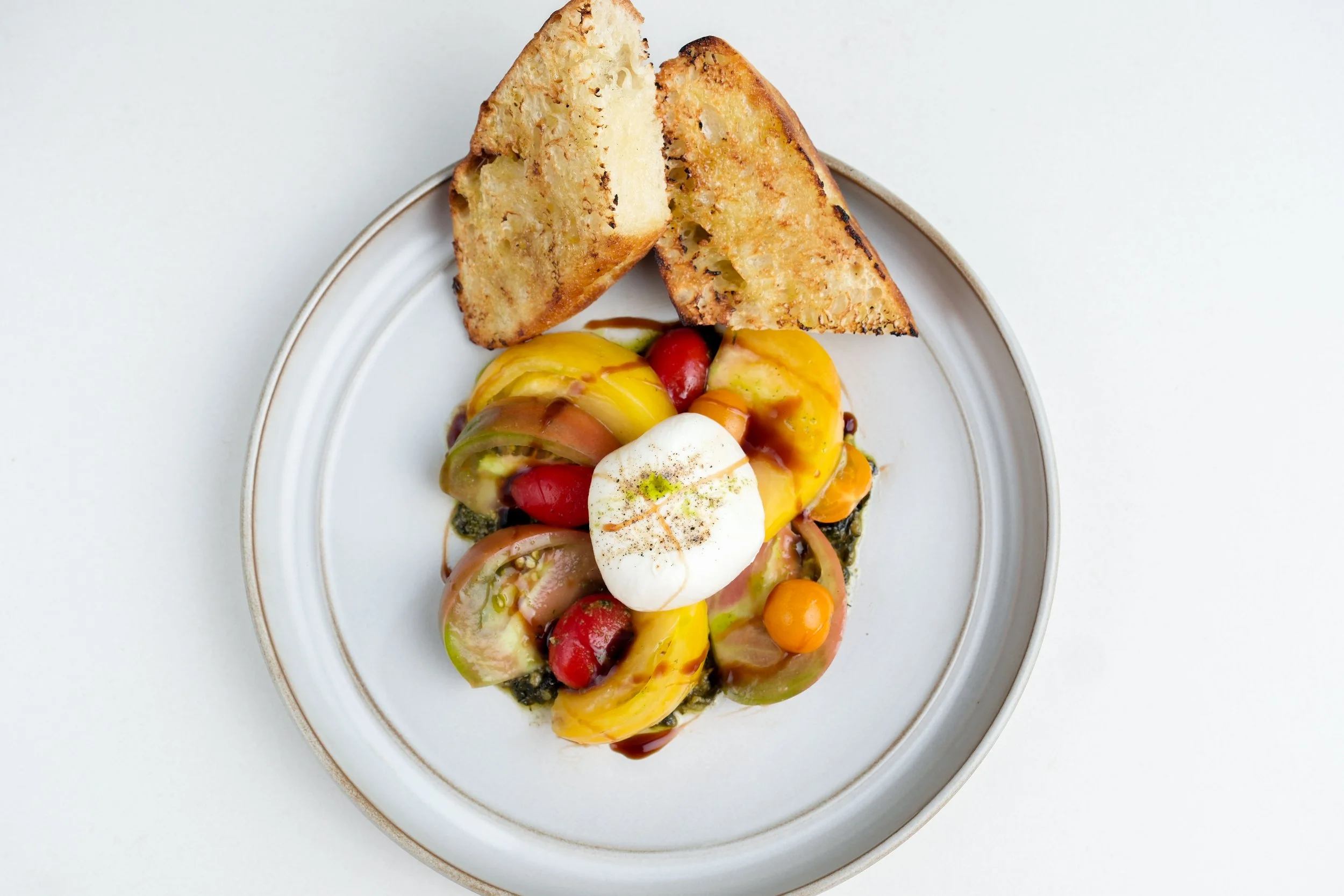 A white plate with rustic toasted bread slices and a colorful heirloom tomato salad topped with a poached egg, drizzled with sauce, on a white surface.