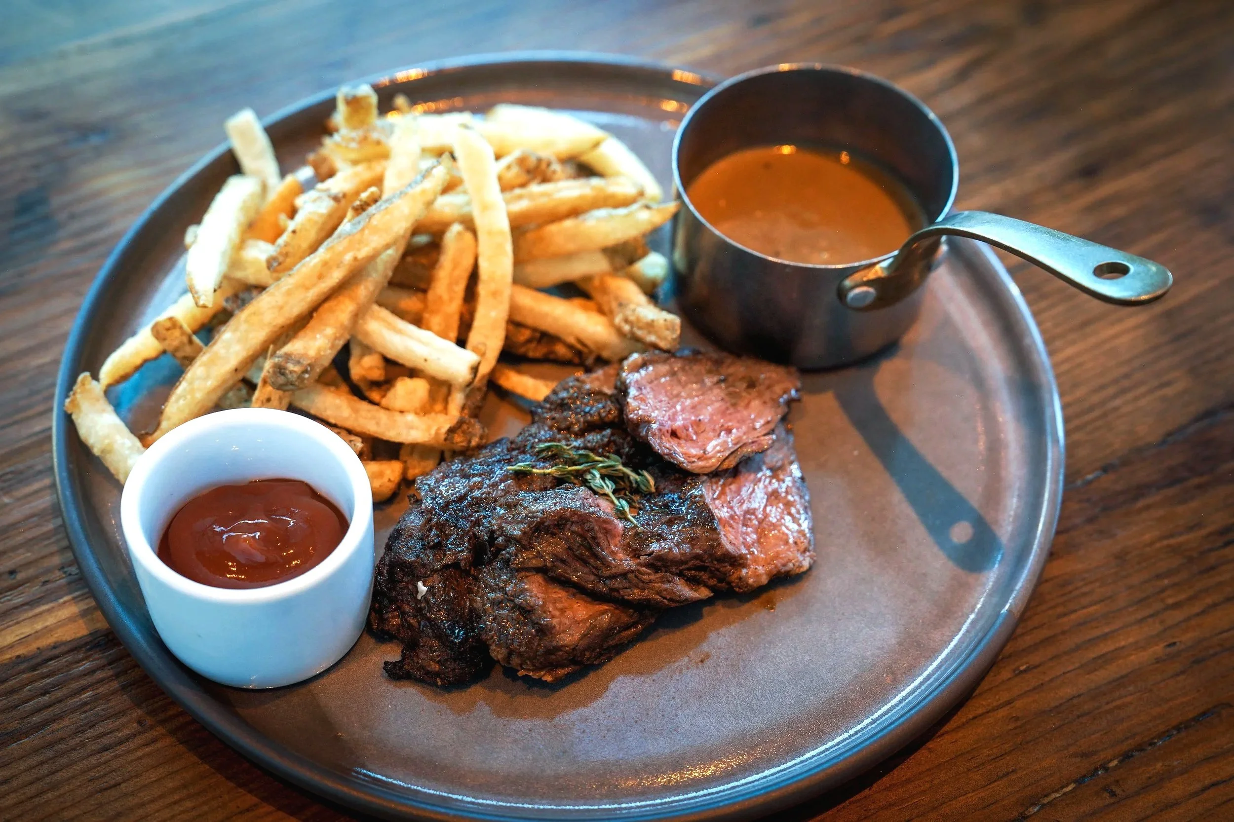 Plate of French fries with a side of dipping sauce, tender roasted beef, and a bowl of brown gravy or sauce, on a wooden table.