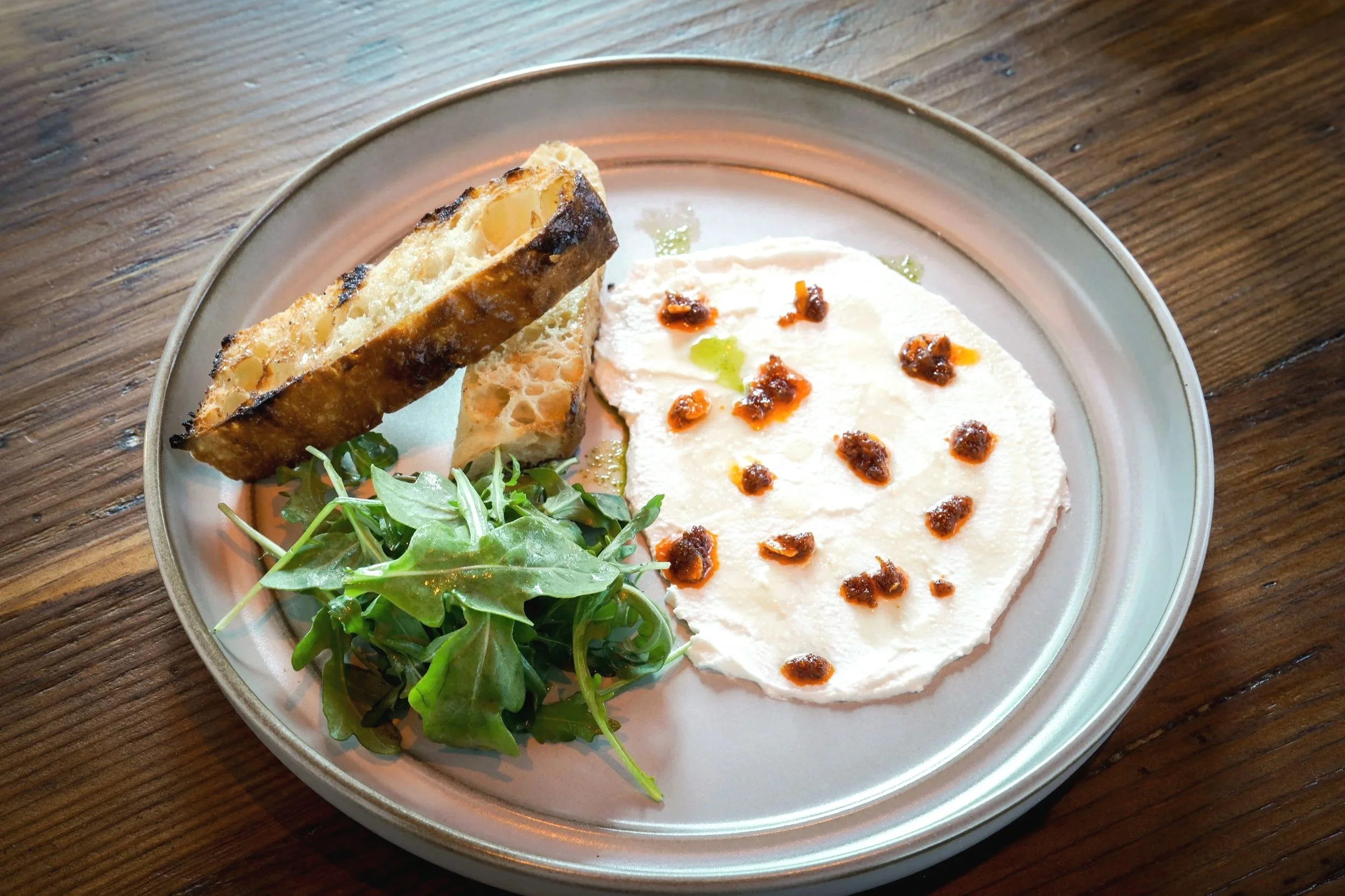 A plate of food with two toasted bread slices, a side of arugula greens, and a serving of creamy white dip topped with red and green sauce.