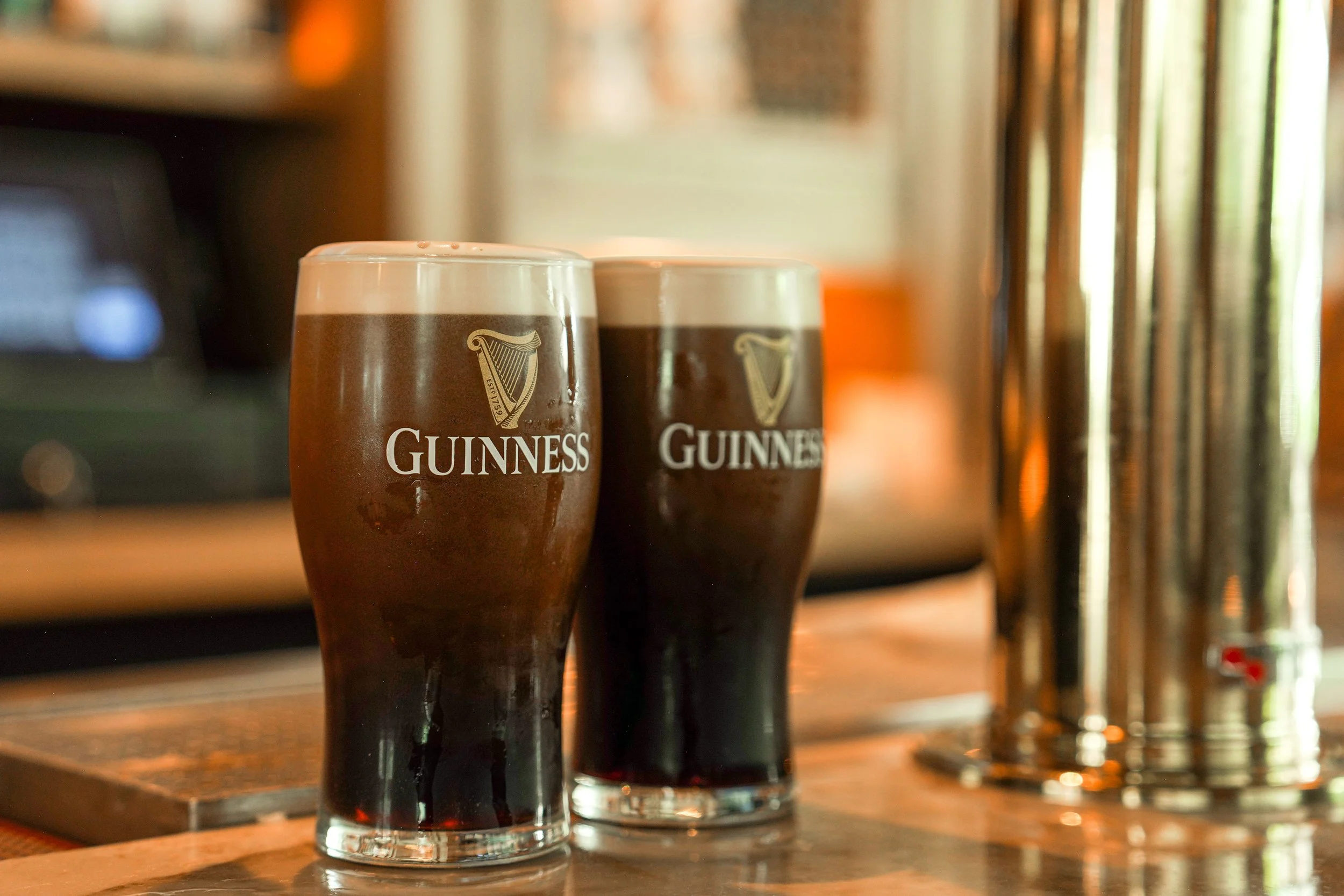 Two glasses of Guinness beer on a wooden bar counter, with one partially visible in the background, and beer taps on the right side of the image.