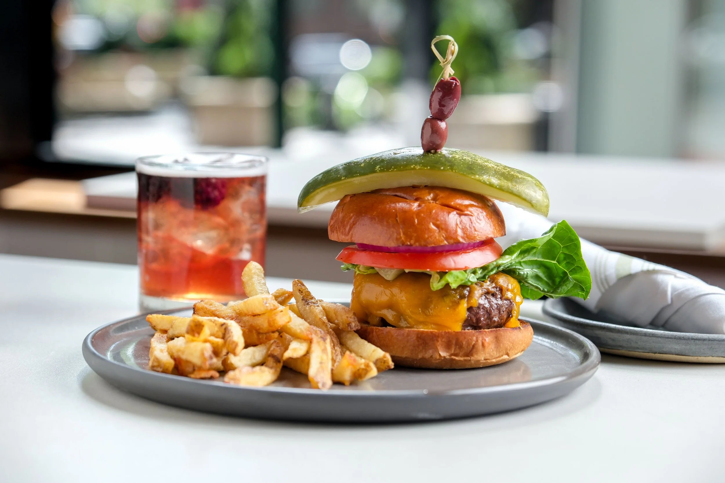 A plate with a hamburger containing lettuce, tomato, onion, pickles, cheese, and a beef patty, topped with a pickle spear and two olives on a toothpick. Next to the burger is a small serving of French fries. In the background, there is a glass of red