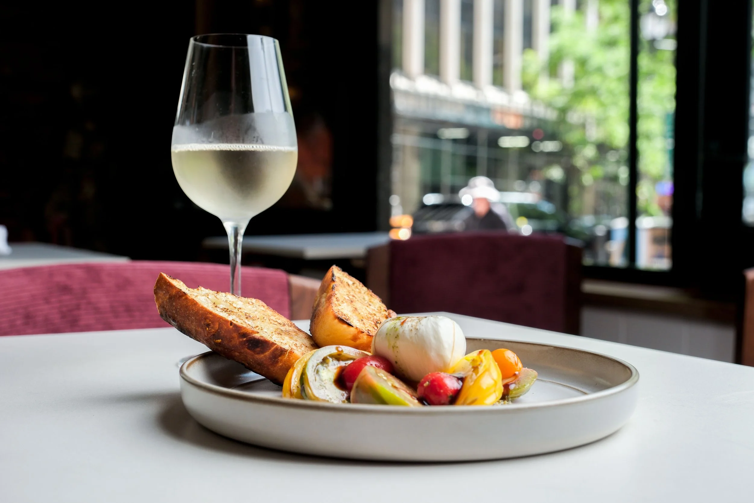 A white plate of colorful salad with vegetables and bread, and a glass of white wine on a table inside a restaurant near a window.