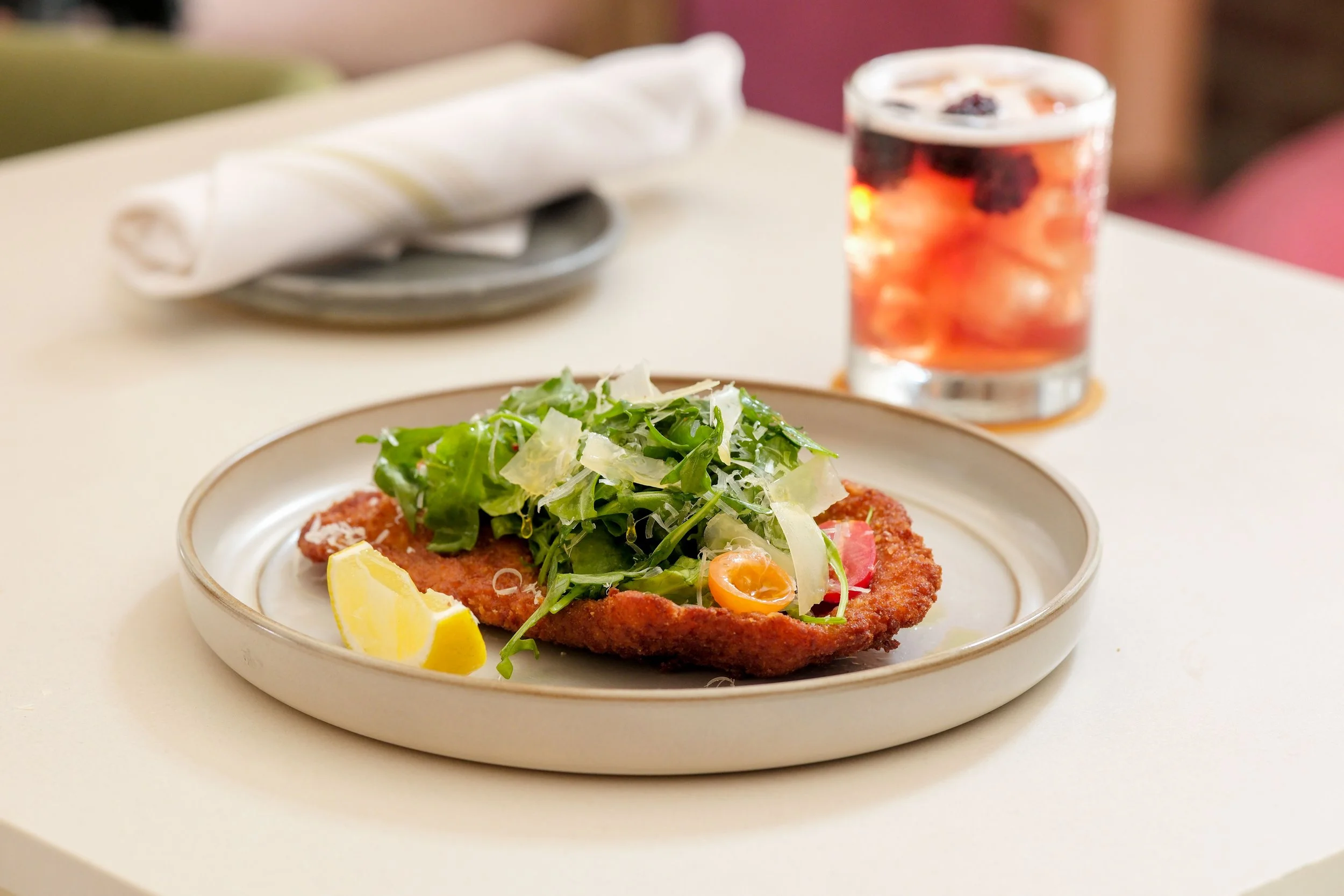 A plate of breaded fish fillet topped with fresh greens and shredded cheese, with lemon wedges on the side. A glass of iced tea with ice and berries is in the background on a white table.