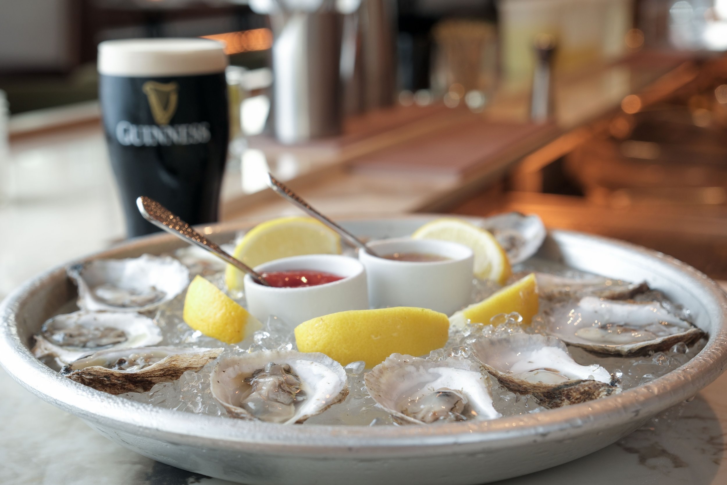 A bowl of fresh oysters on ice garnished with lemon wedges, accompanied by small cups of cocktail sauce and horseradish, with a pint of Guinness beer in the background inside a restaurant or pub.