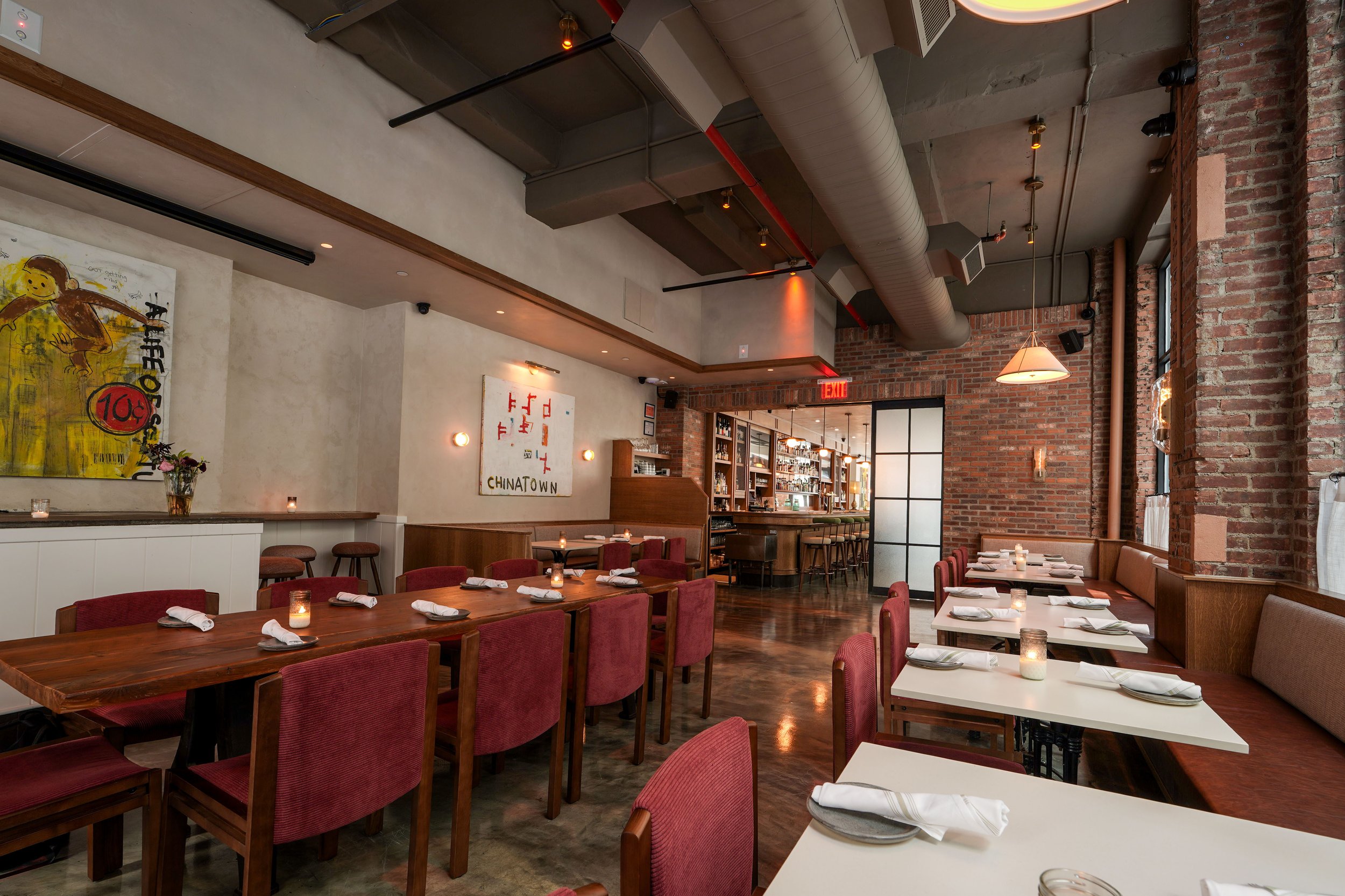 Interior of a modern restaurant with exposed brick walls, wooden accents, and dim lighting. The dining area has tables set with plates, napkins, and flickering candles, with artwork on the walls and a bar area in the background.