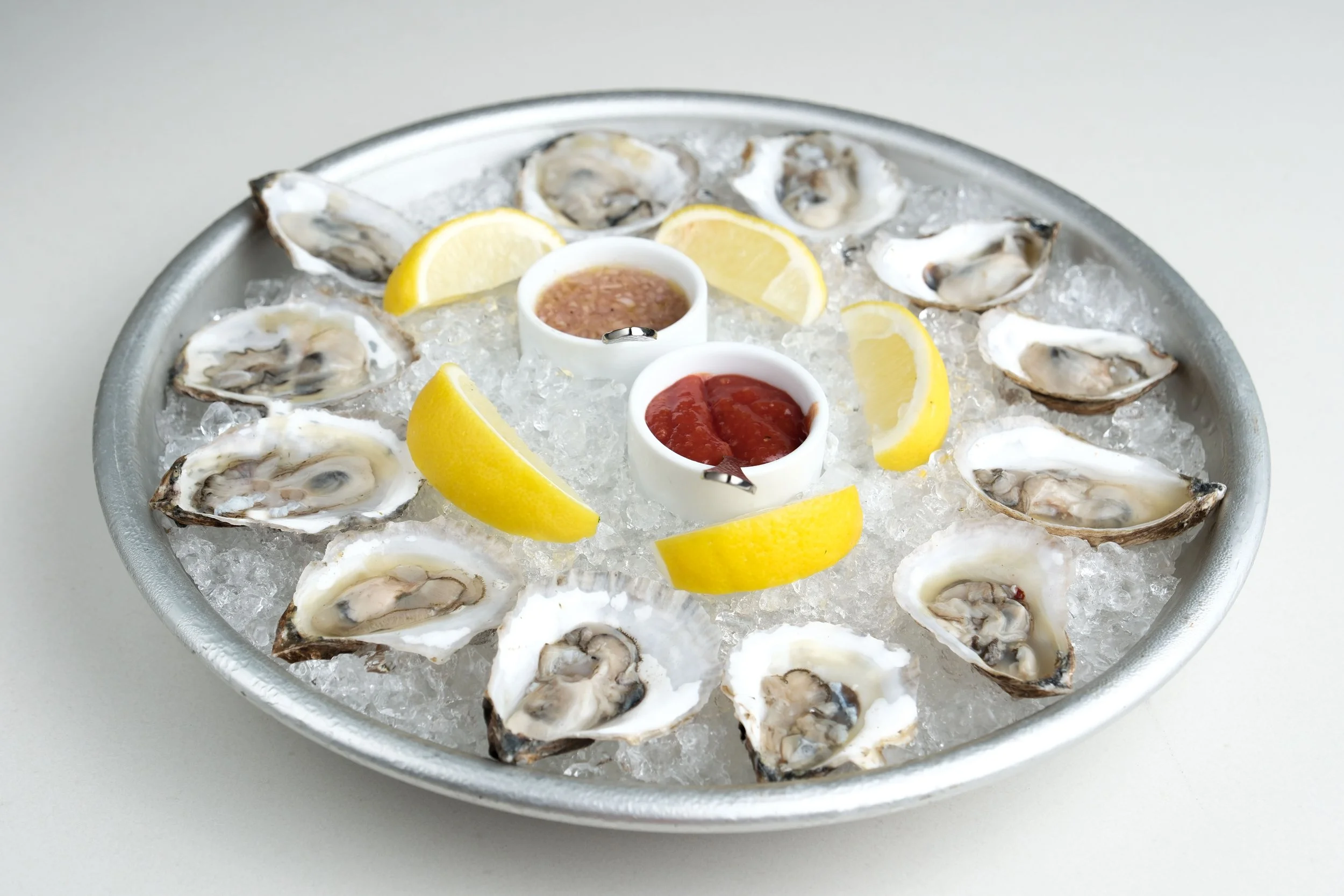 A metal tray of fresh oysters on ice, with lemon wedges and two small containers of cocktail sauce and hot sauce.