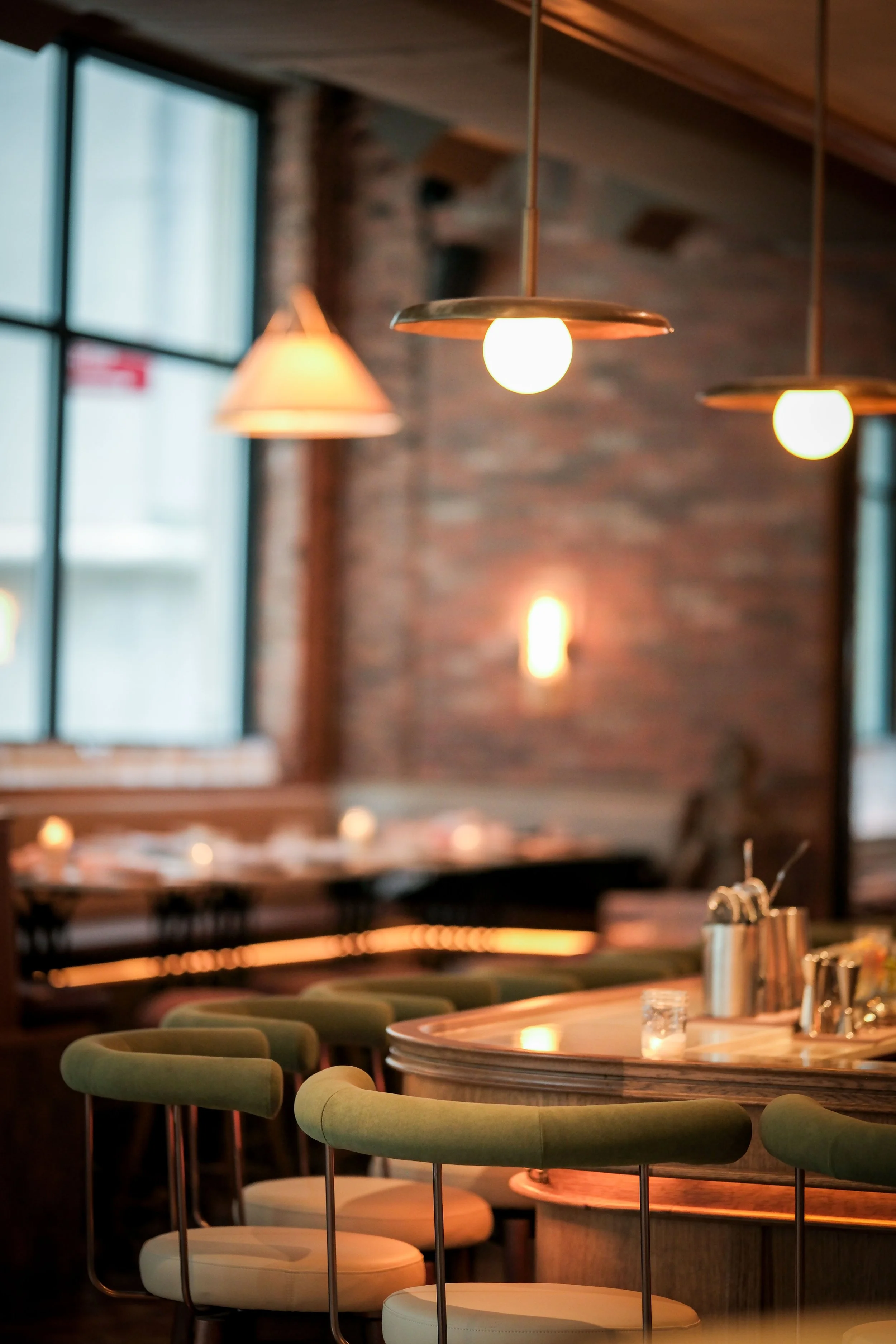 Interior of a restaurant or cafe with hanging pendant lights, a large window, brick wall, and a round table with chairs.