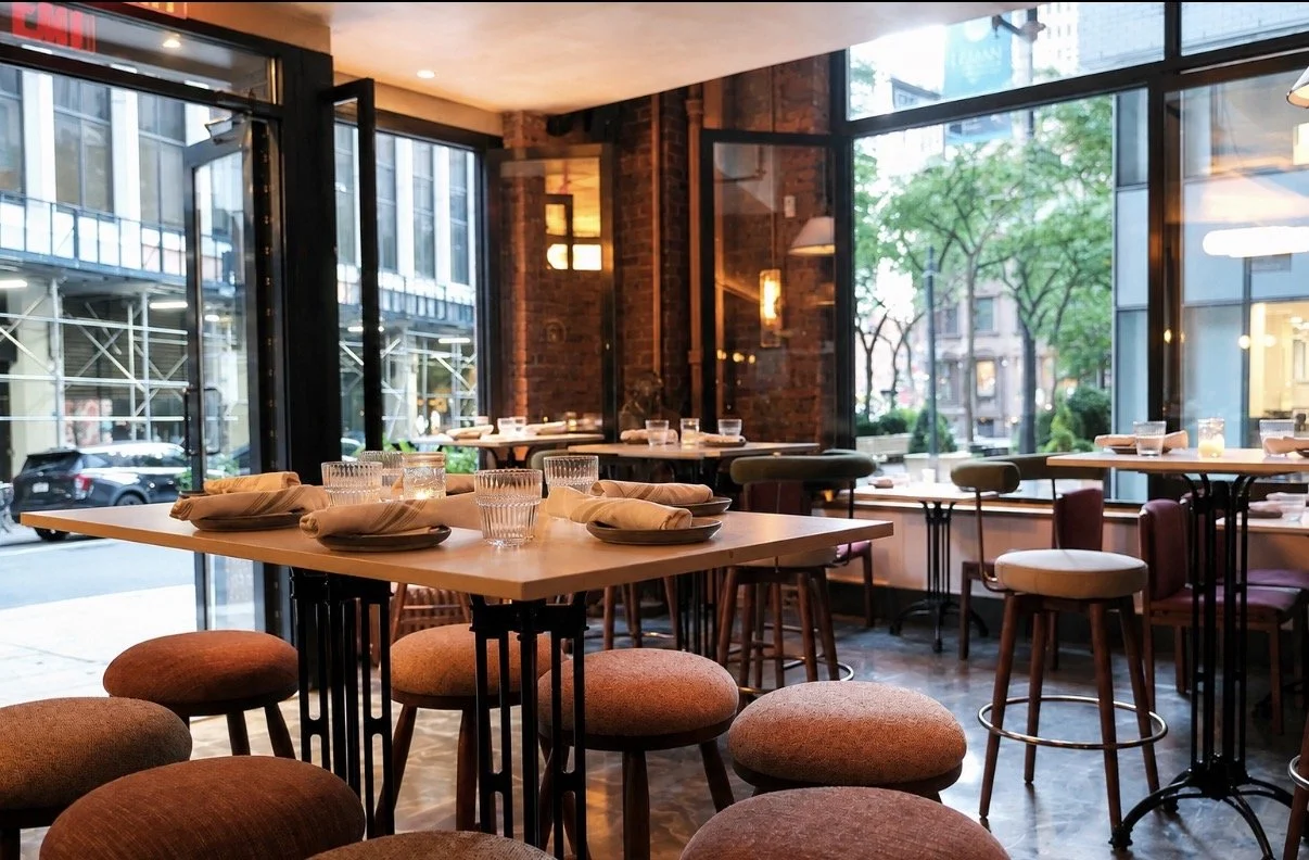 Interior of a modern restaurant with wooden tables set with napkins, glasses, and plates, surrounded by stools and chairs, large windows revealing a city street outside, trees, and buildings.