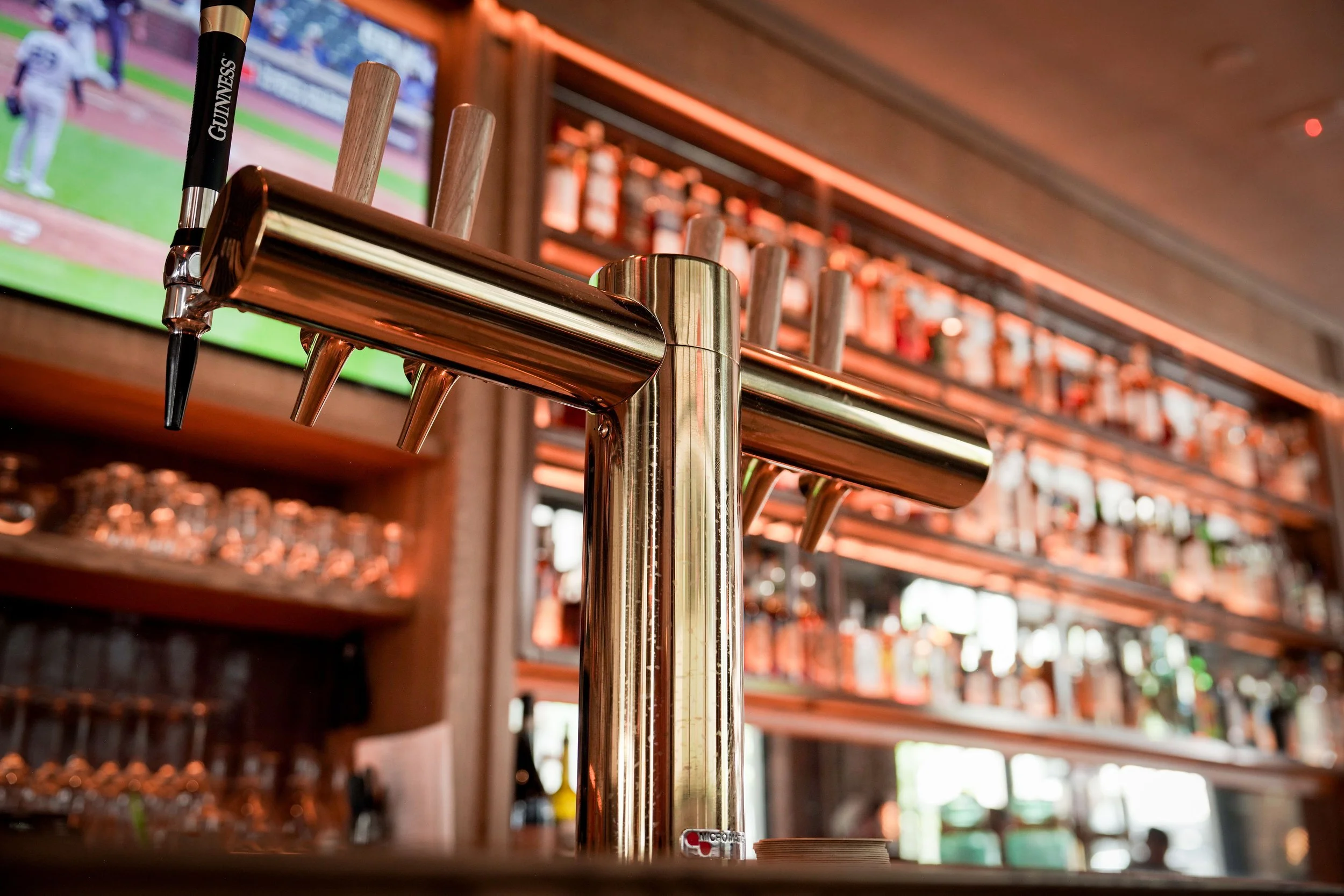 Close-up of beer tap handles mounted on a metallic draft tower, with a bar or restaurant interior blurred in the background.