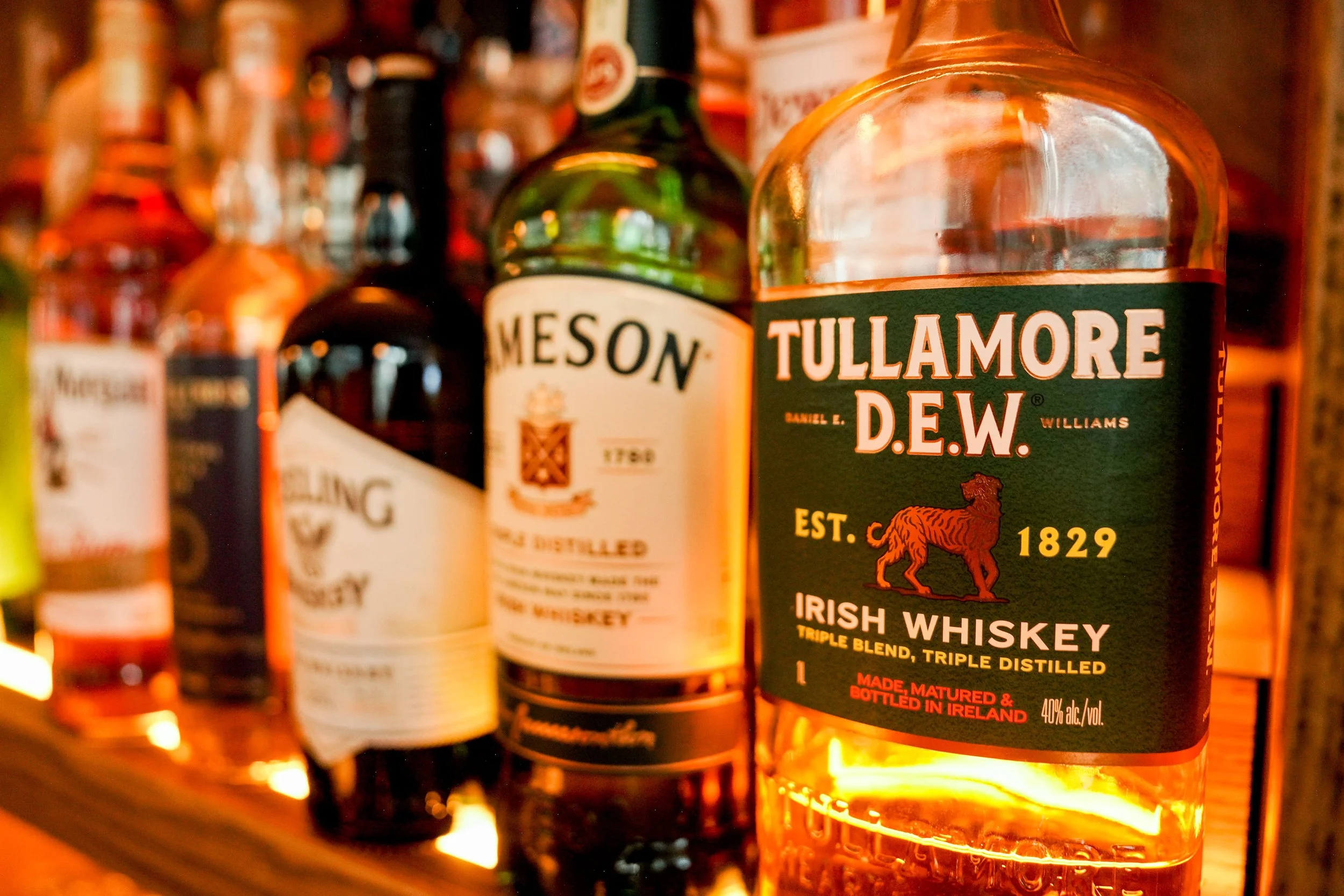 A shelf of whiskey bottles, with the prominent bottle labeled Tullamore D.E.W., Irish whiskey, triple blend, triple distilled, established 1829.