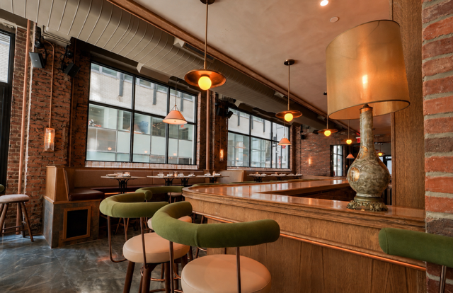 Interior of a modern restaurant with large windows, brick walls, hanging pendant lights, a wooden bar counter, and green upholstered chairs.