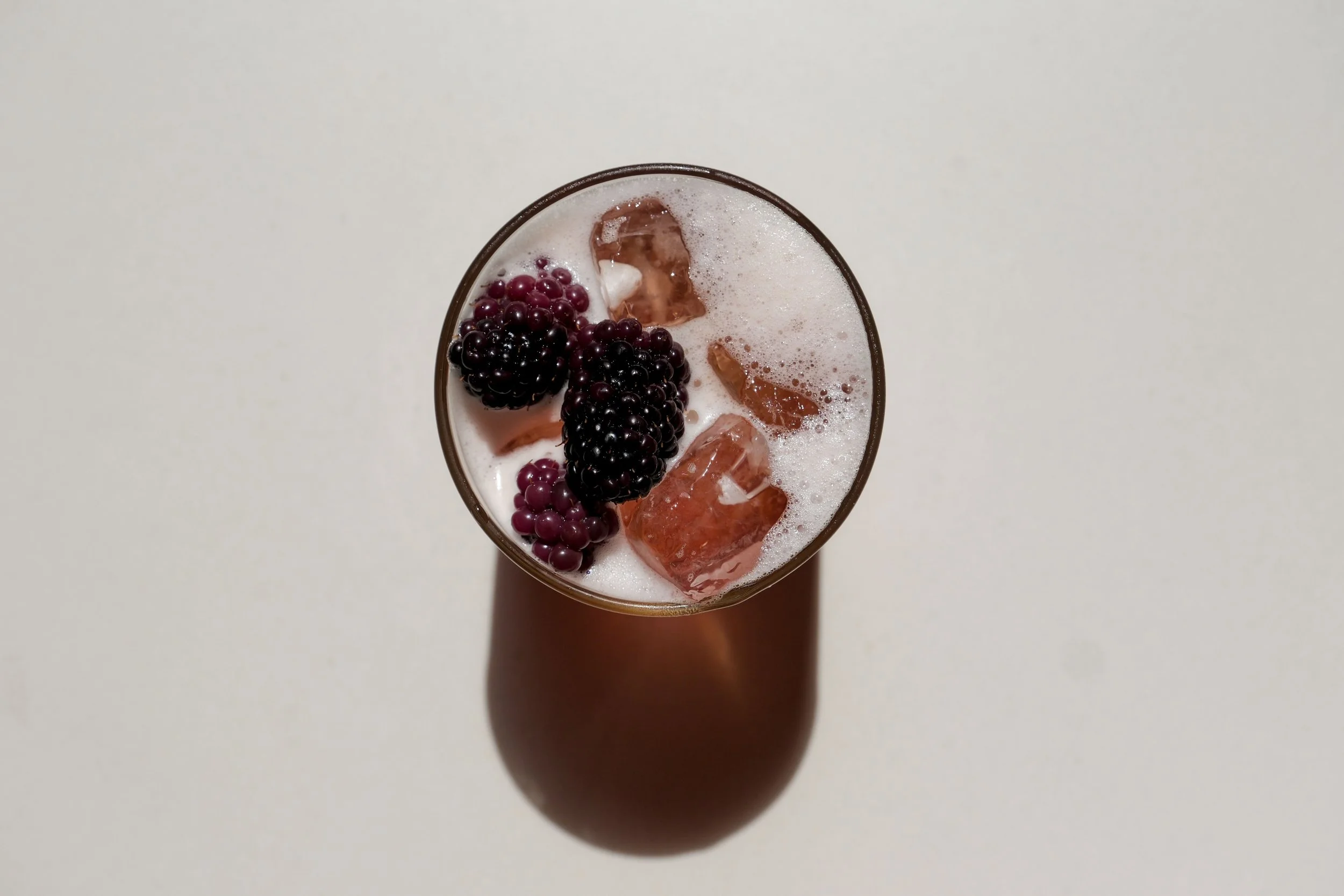 A top-down view of a glass of mixed berry cocktail with ice, blackberries, and foam, set against a white background.