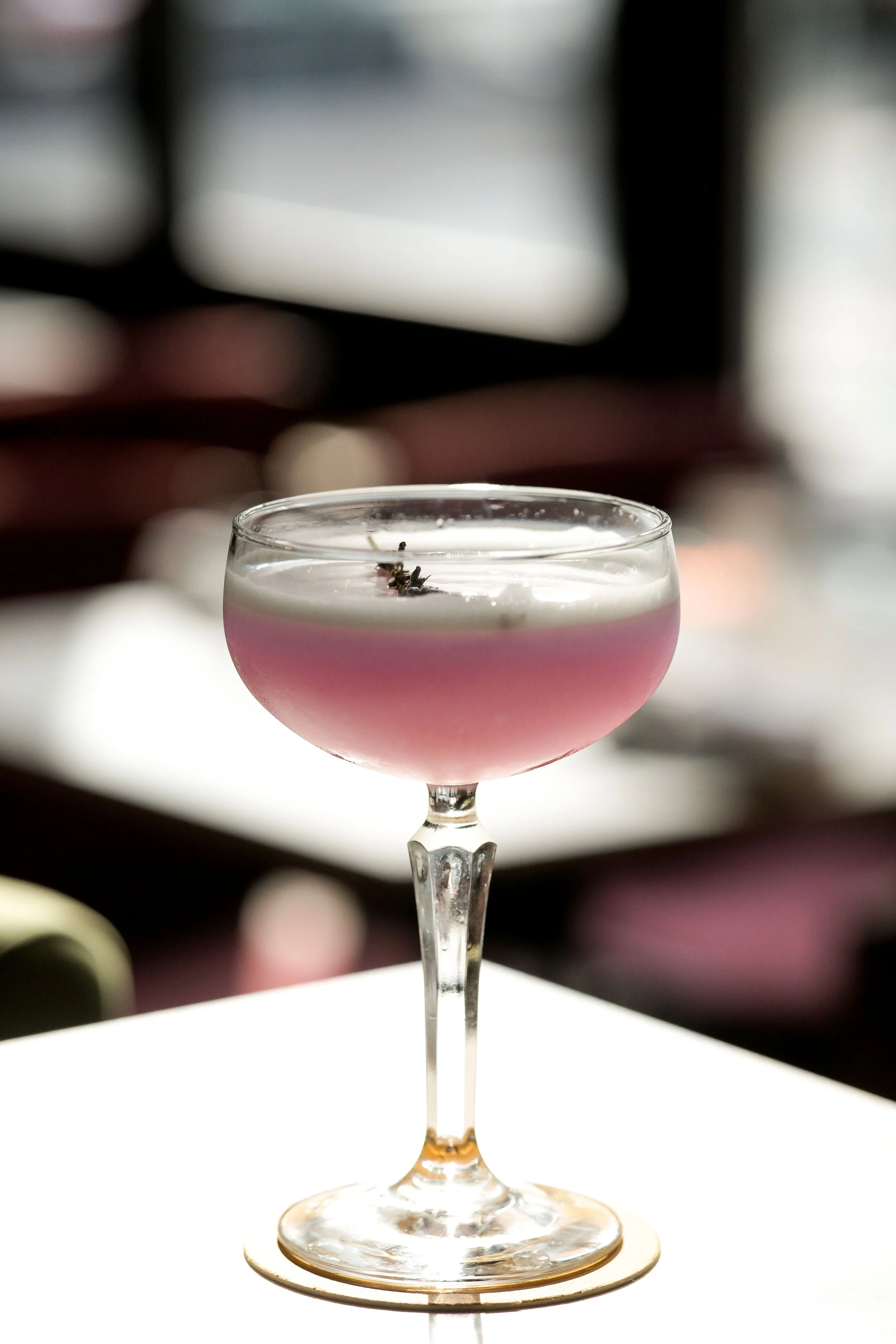 A cocktail in a coupe glass with a pink hue and a small garnish on top, set against a blurred background with white and black elements.