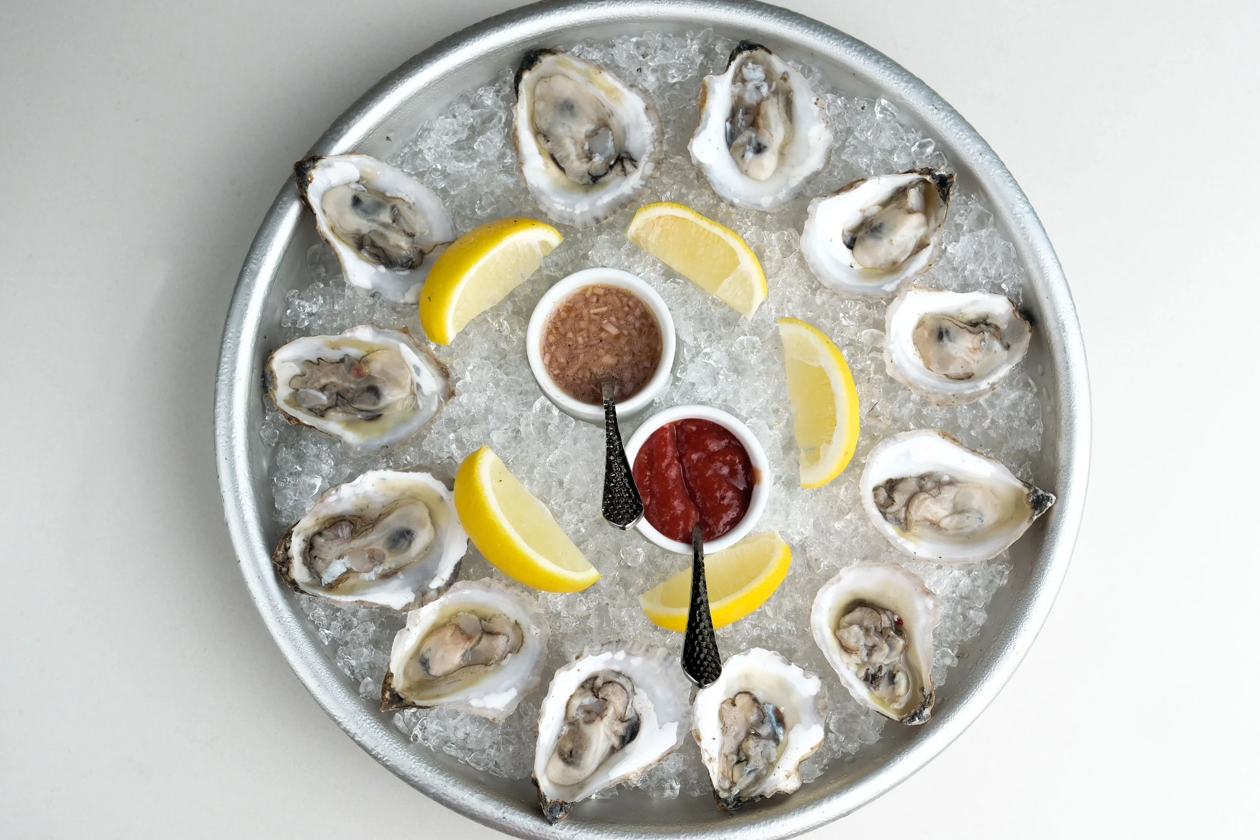 A round tray filled with crushed ice containing fresh oysters on the half shell, lemon wedges, and two small bowls of sauce, one with cocktail sauce and the other with a tartar or mignonette sauce.