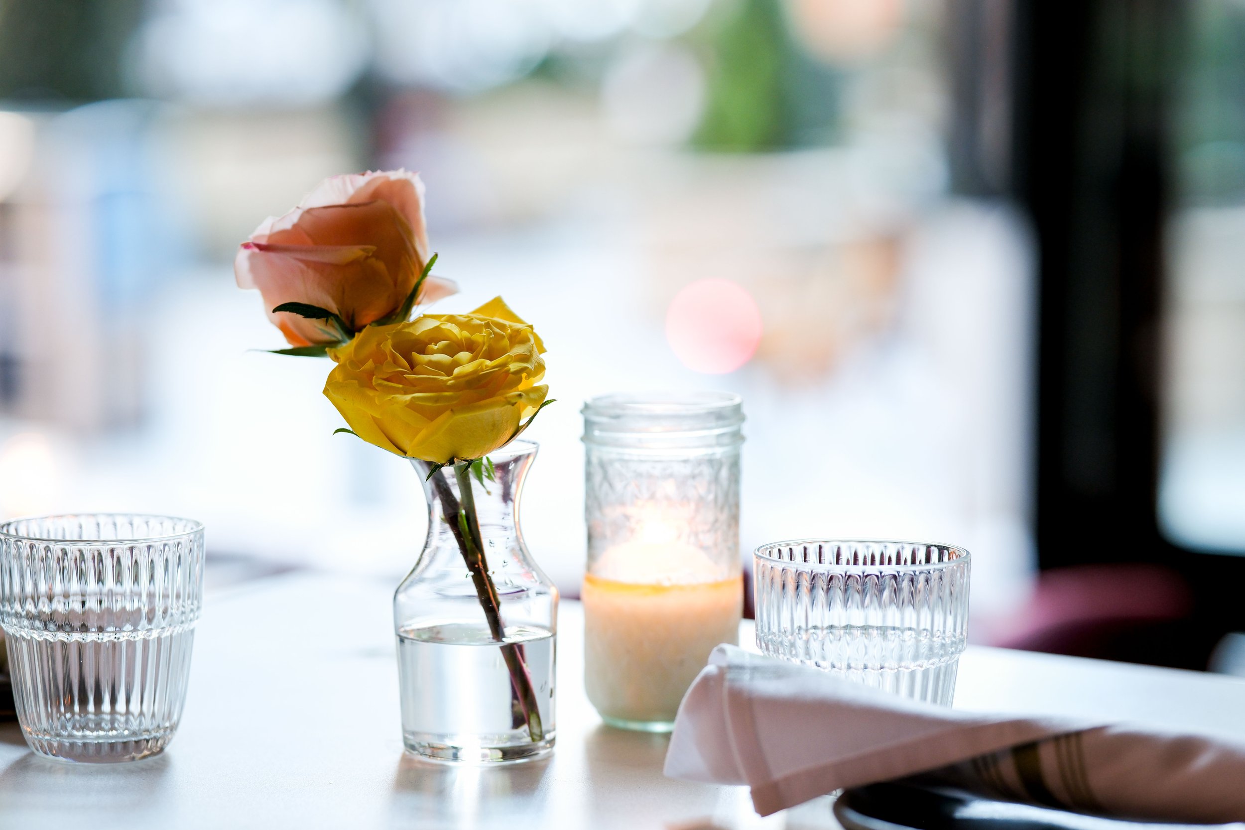 A table centerpiece with pink and yellow roses in a clear glass vase, surrounded by clear ribbed glasses and a candle, with a blurred background.