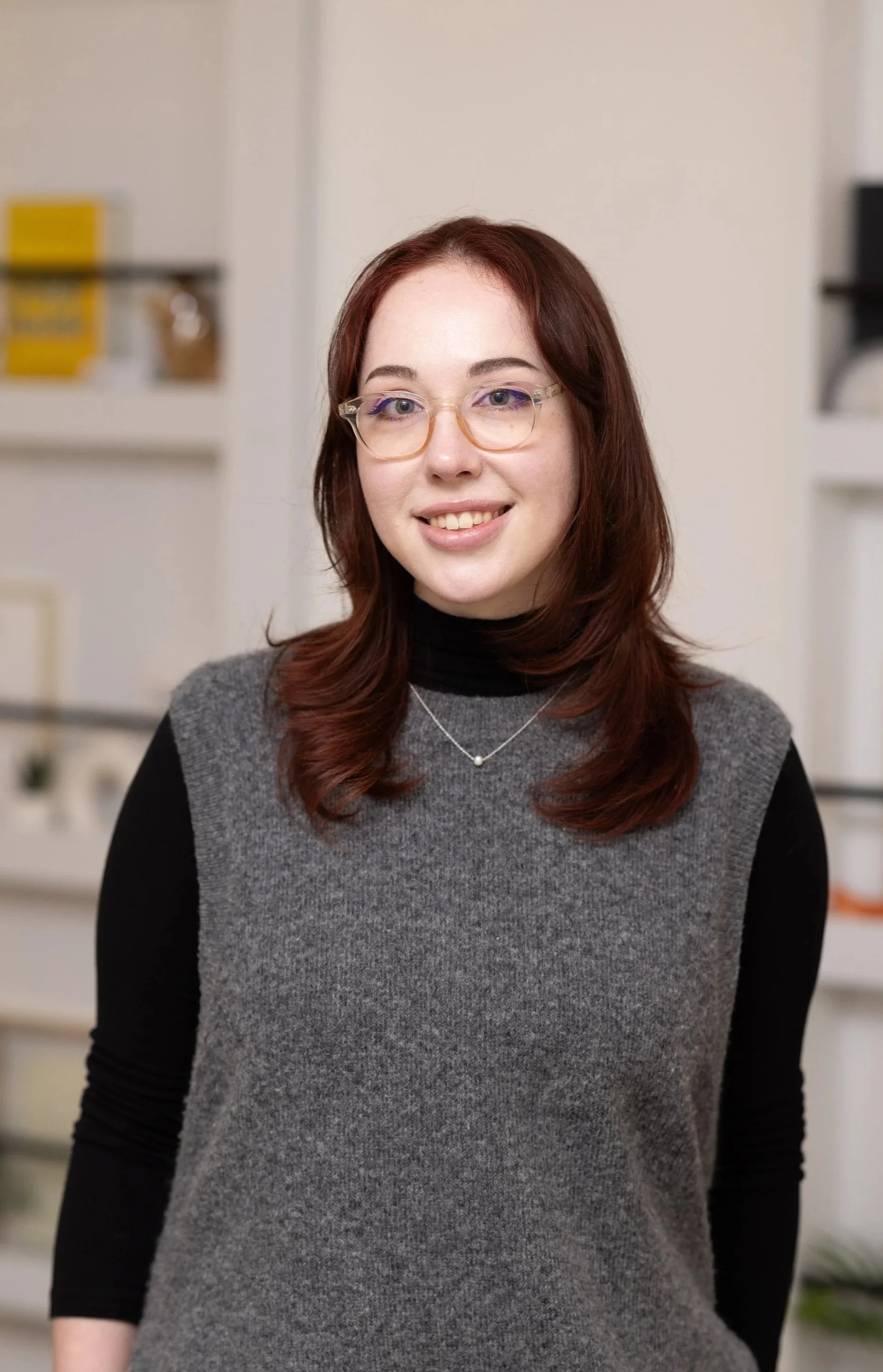A young woman with red hair, glasses, and a gray and black sweater standing indoors in front of shelves.