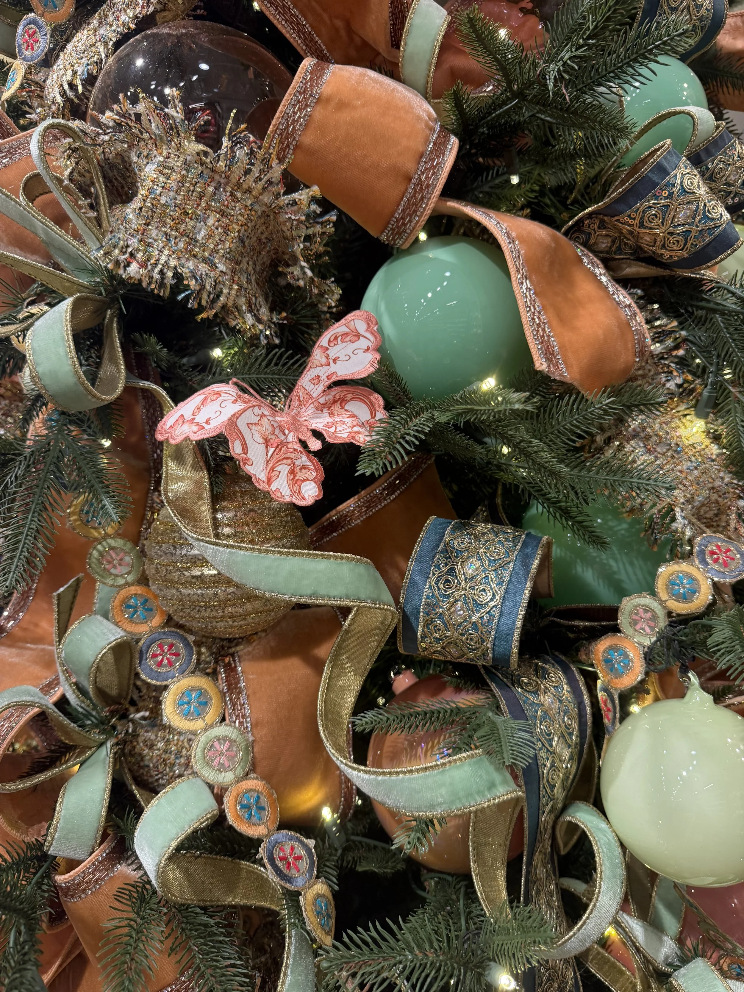 Close-up of Christmas decorations featuring green pine branches, colorful ribbons, embroidered ornaments, and large pastel green baubles.