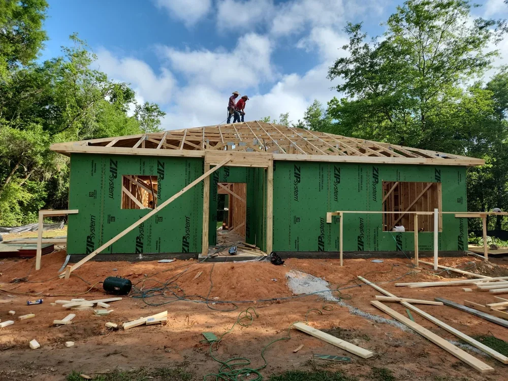 Under construction house with green sheathing, wooden framing on the roof, and two workers on top working on the roof. The area around the house is dirt with scattered building materials and tools.