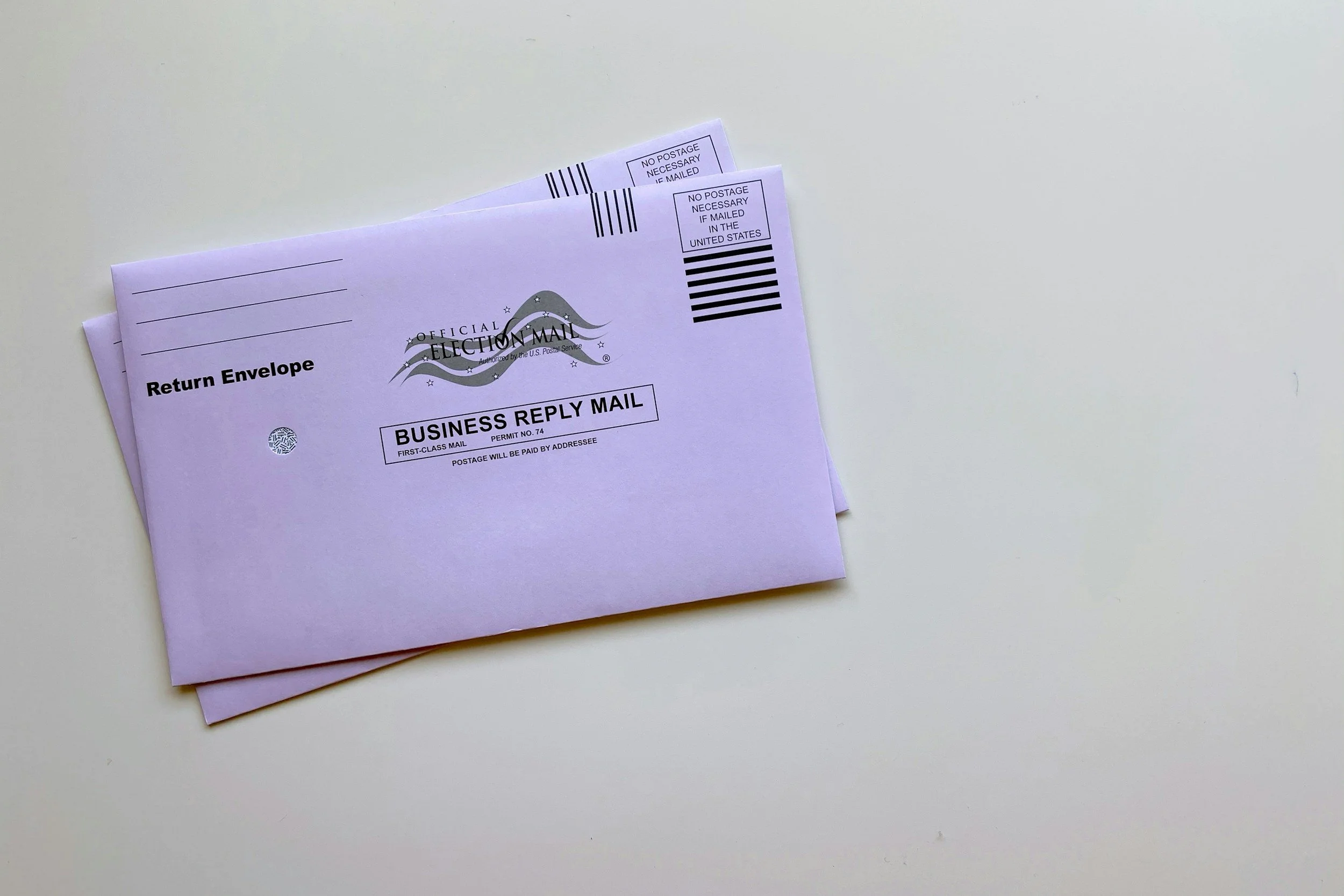 Two pink envelopes with barcode labels and 'Official Election Mail' markings, placed on a white surface.