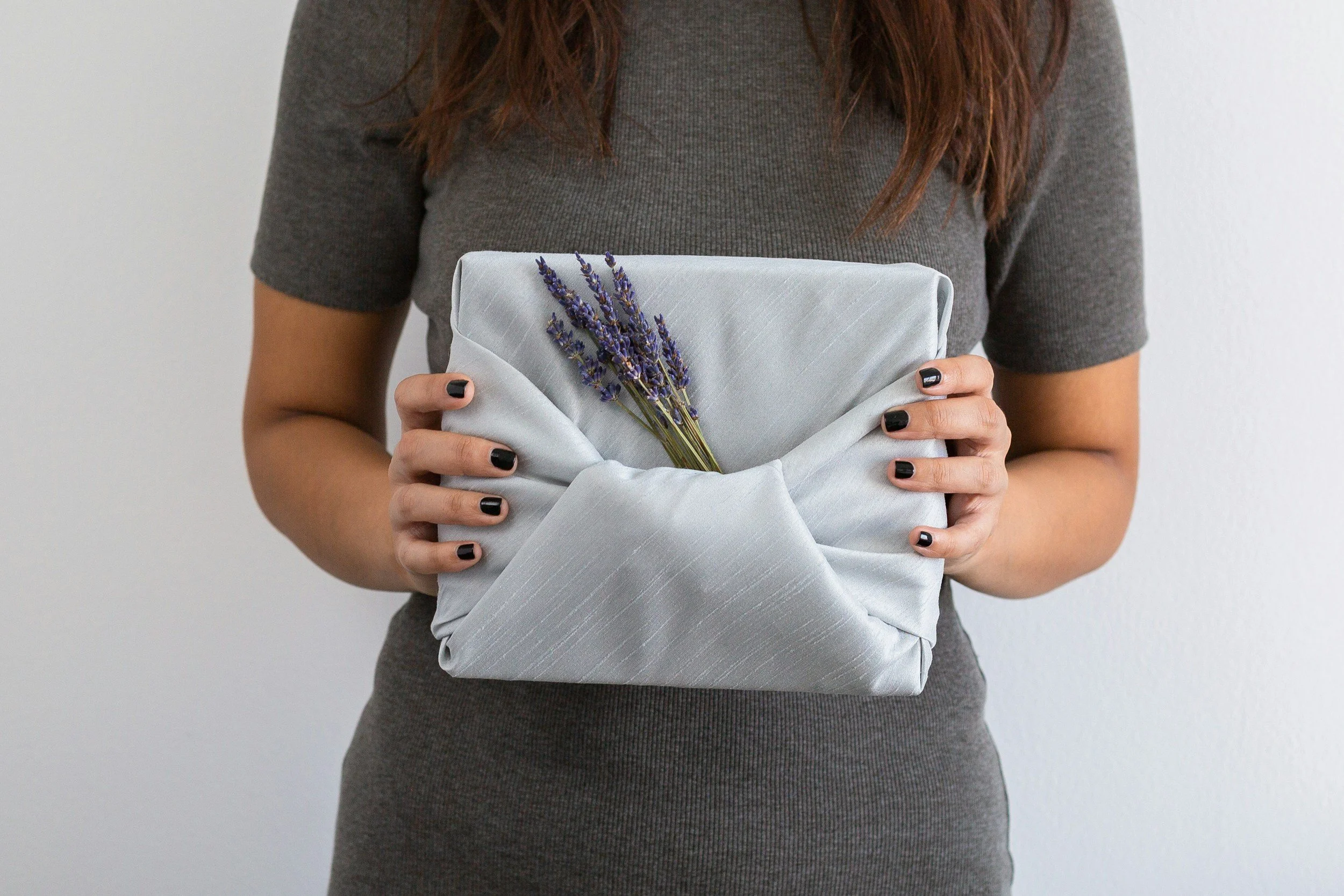 A woman in a gray dress holds a wrapped gift with a small lavender bouquet attached.
